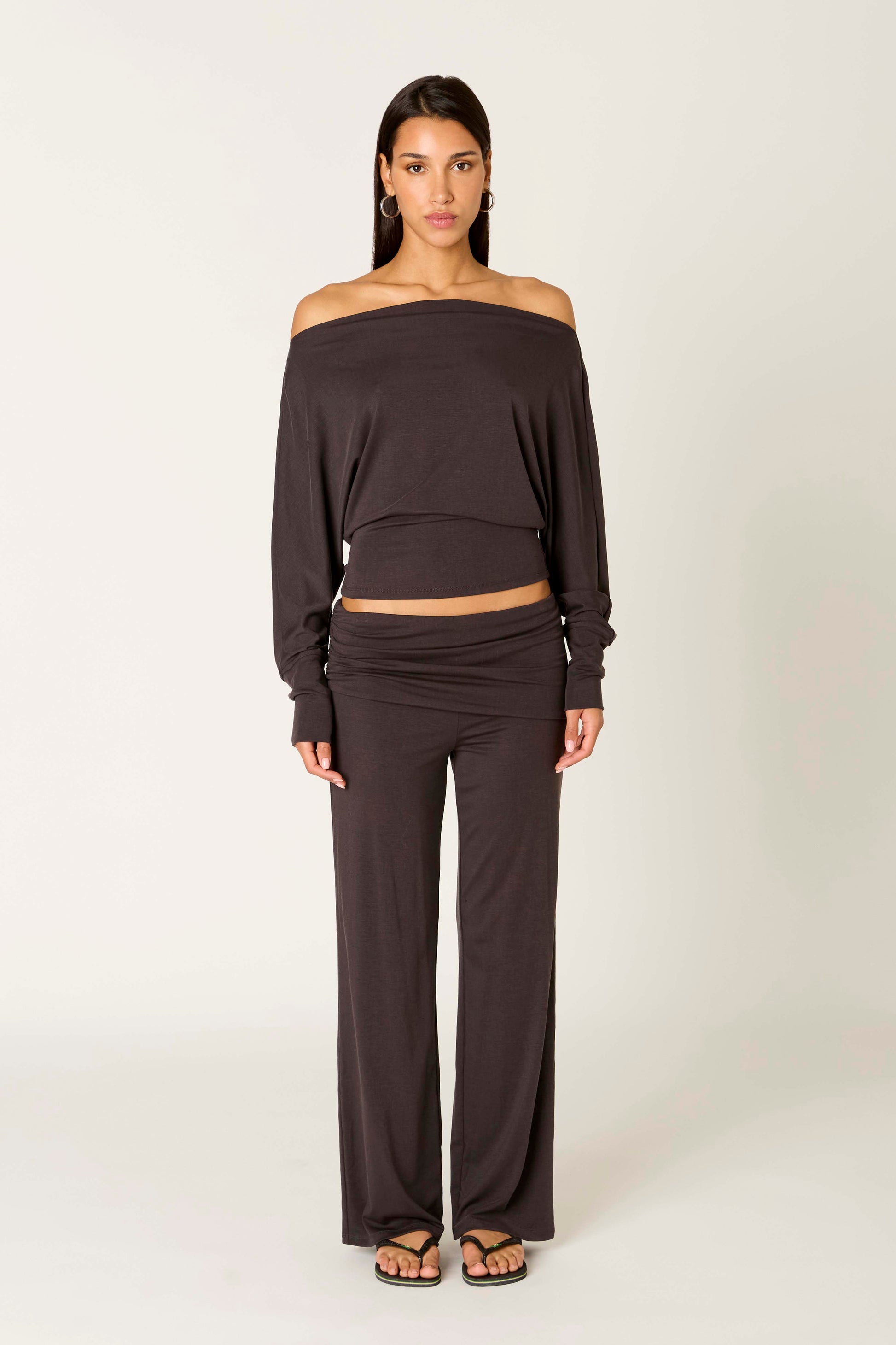 A woman faces forward against a plain light background, wearing the Nia Elisabetta Top—an off-the-shoulder long-sleeve black blouse with a blouson silhouette—paired with matching black pants and flip-flops.