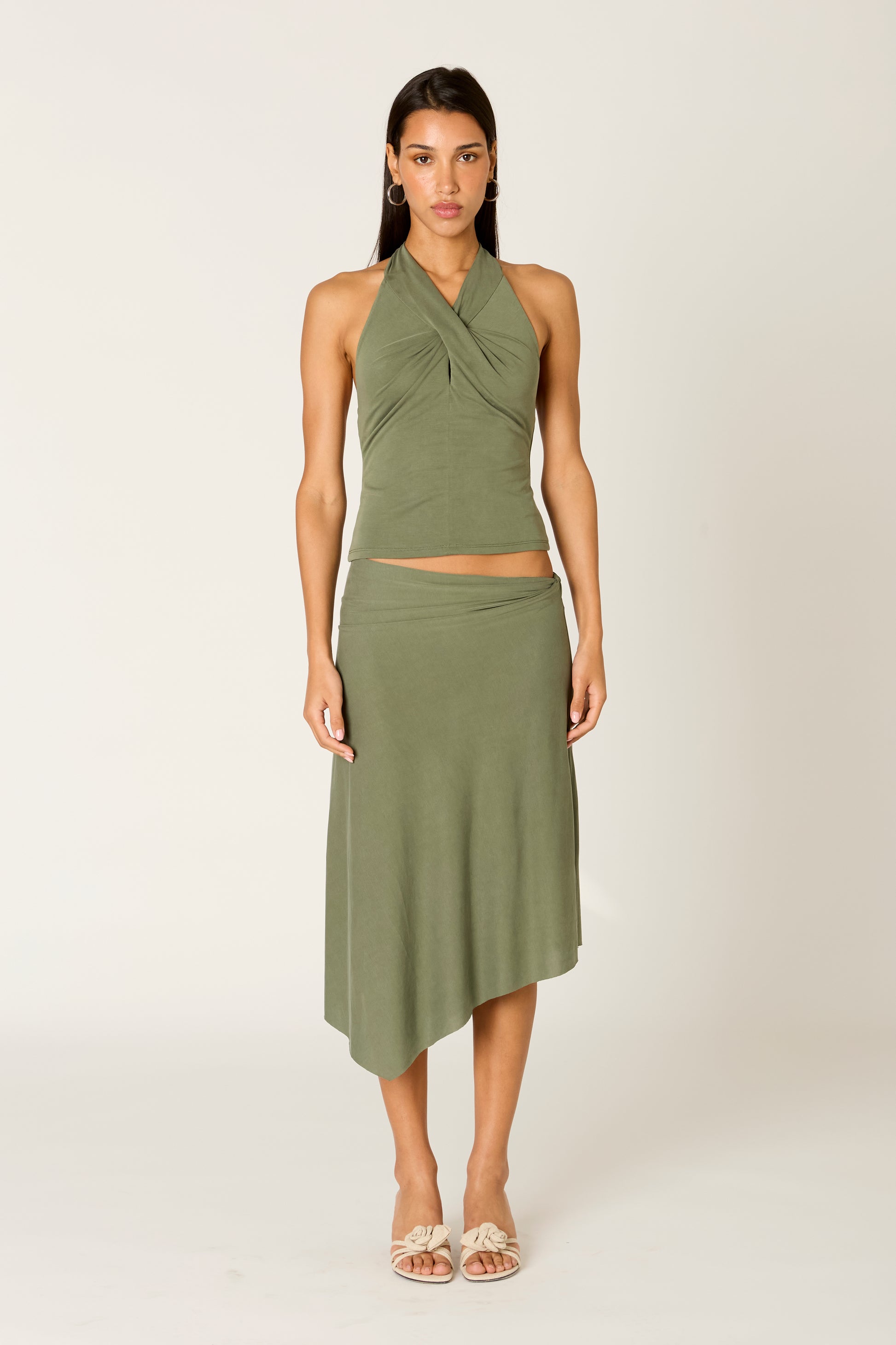 A woman stands against a plain light background, wearing Nia’s Francesca Skirt—a sleeveless, olive green halter top paired with the matching asymmetrical midi skirt—styled with light-colored sandals and a neutral expression.
