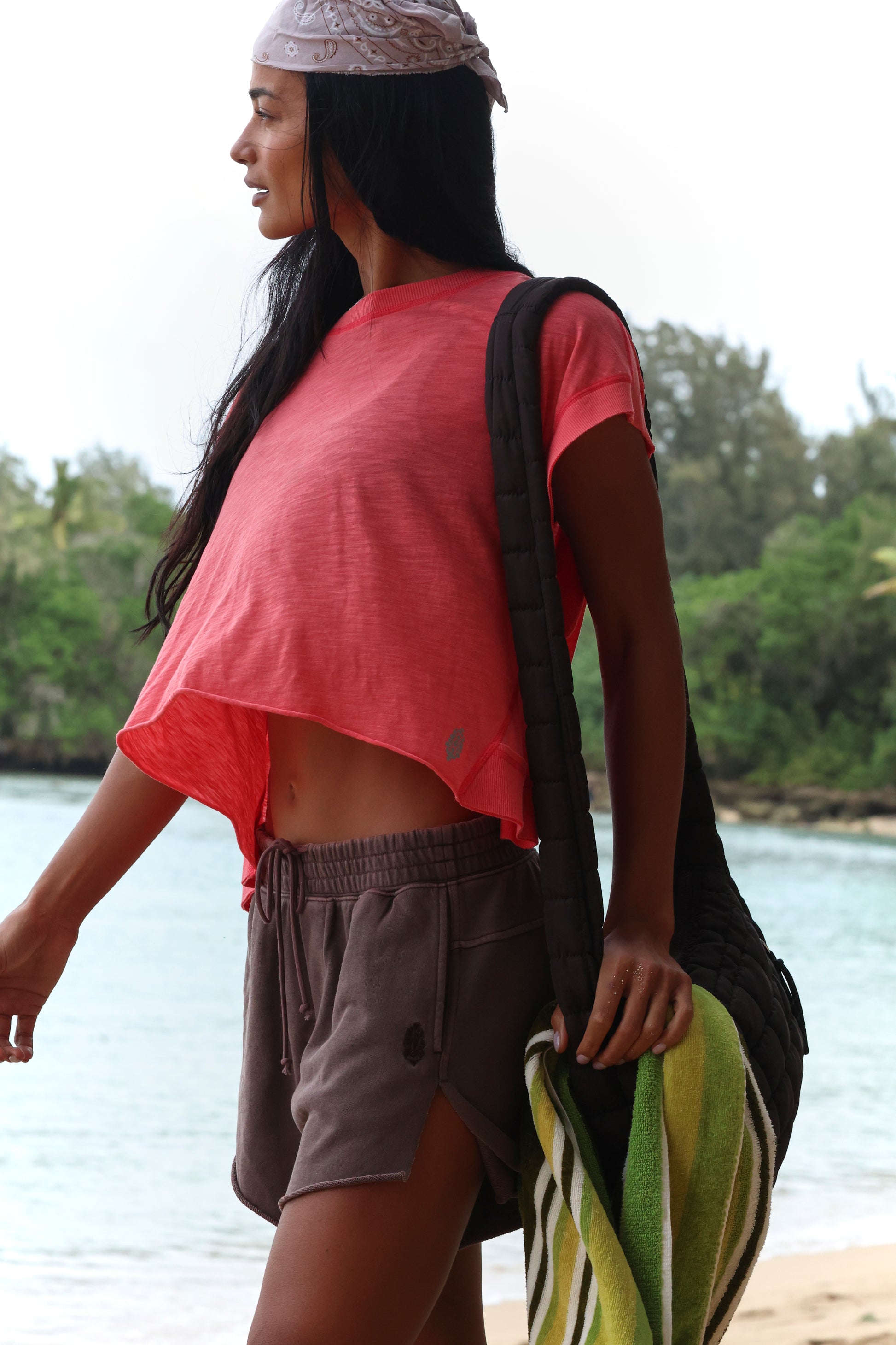 A woman with long dark hair stands by the beach in FP Movement's My Time Tee, a pink asymmetrical crop top with a raw hemline, paired with gray shorts. She holds a striped towel and quilted bag, framed by trees and water in the background.