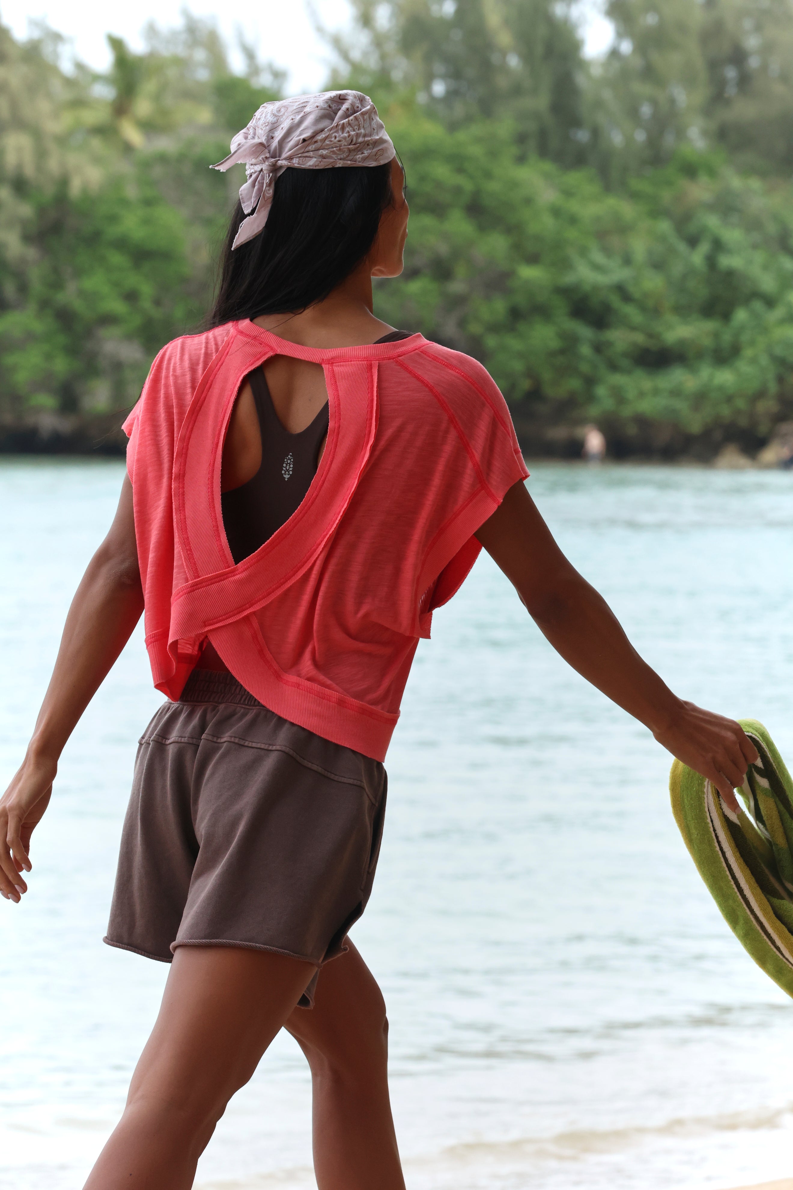 A person with long dark hair wears the FP Movement My Time Tee in pink, paired with brown shorts and a light bandana, as they walk along a beach holding a green towel. Trees and water can be seen in the background.