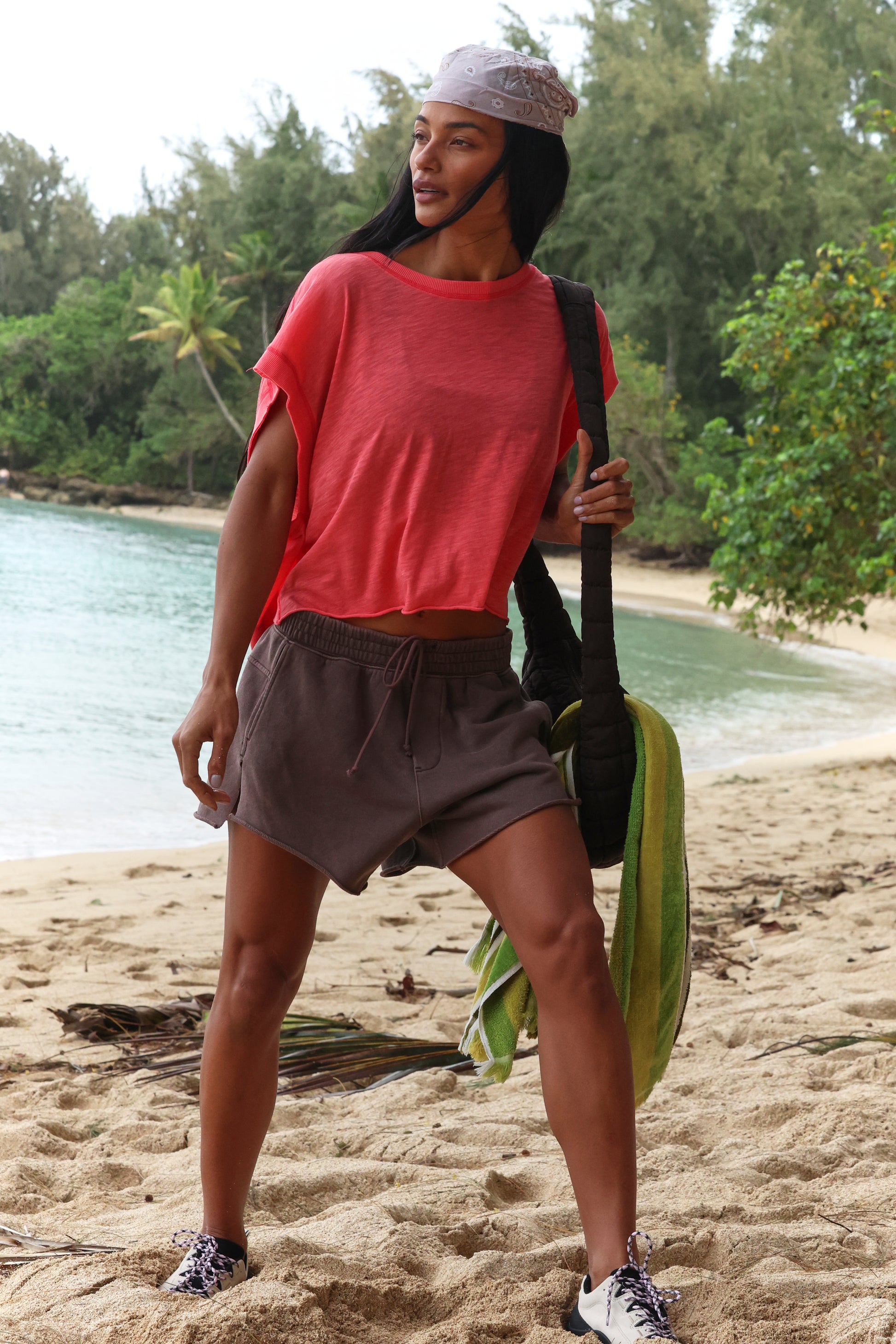 Wearing the FP Movement My Time Tee in red, gray shorts, and sneakers, a person stands on a sandy beach holding a green towel and bag, with trees and water visible in the background.