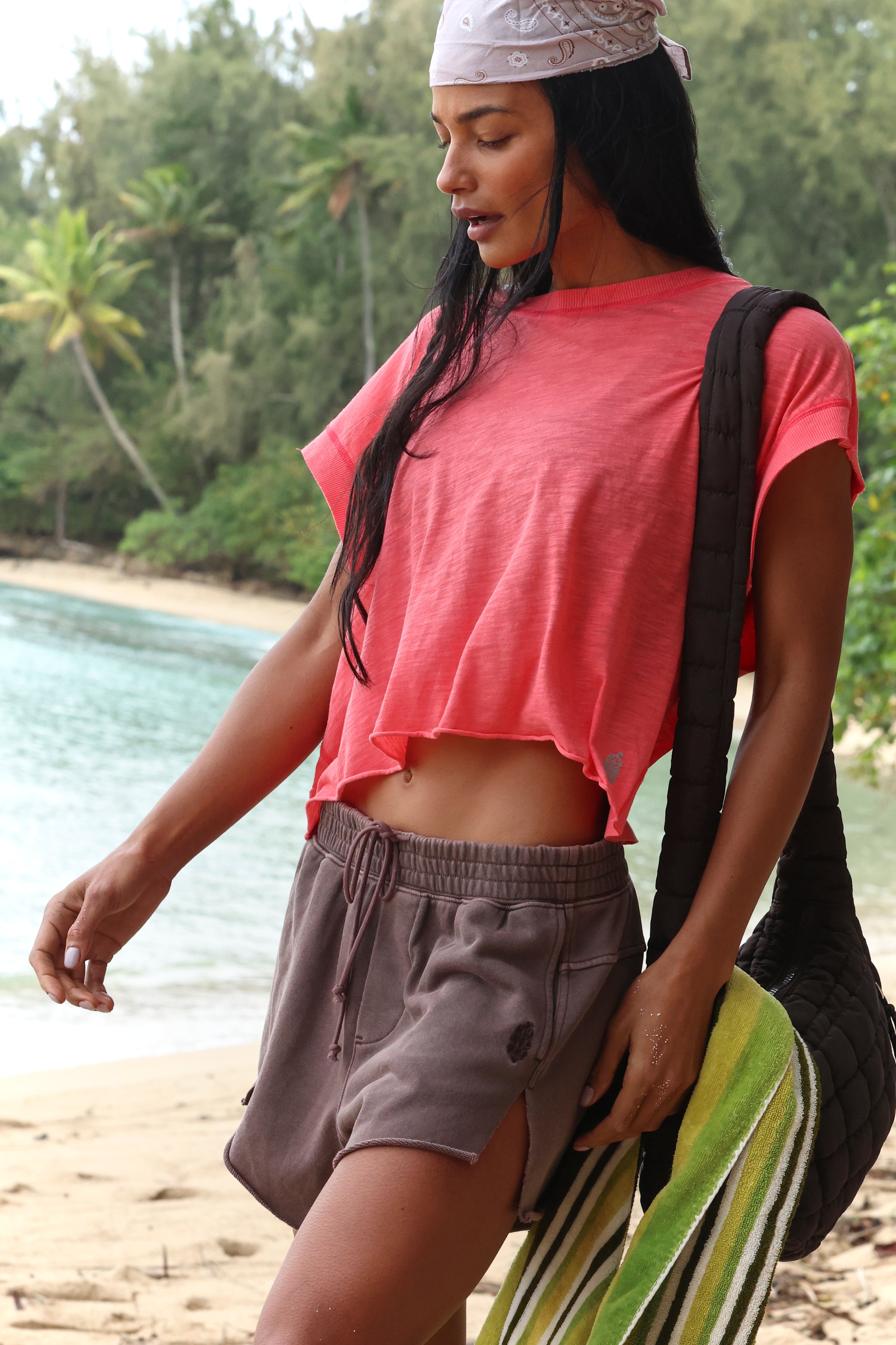 A woman wears the FP Movement My Time Tee in coral, gray shorts, and a light bandana as she stands on a tropical beach holding a striped green towel, with palm trees and the ocean in the background.