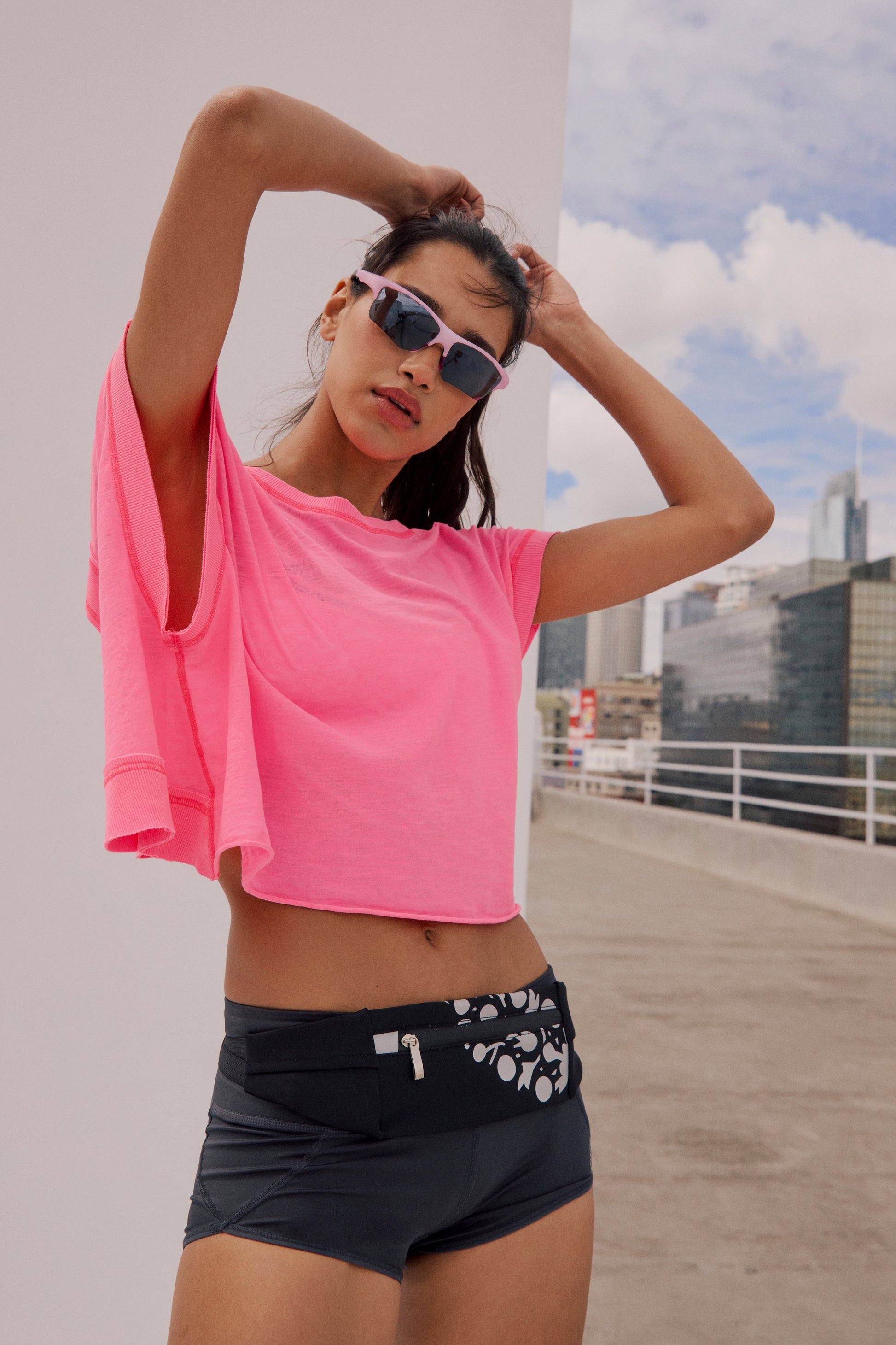 A woman stands on a rooftop with buildings and a cloudy sky, hands in her hair, wearing FP Movement’s My Time Tee in bright pink, sunglasses, black shorts, and a patterned fanny pack.