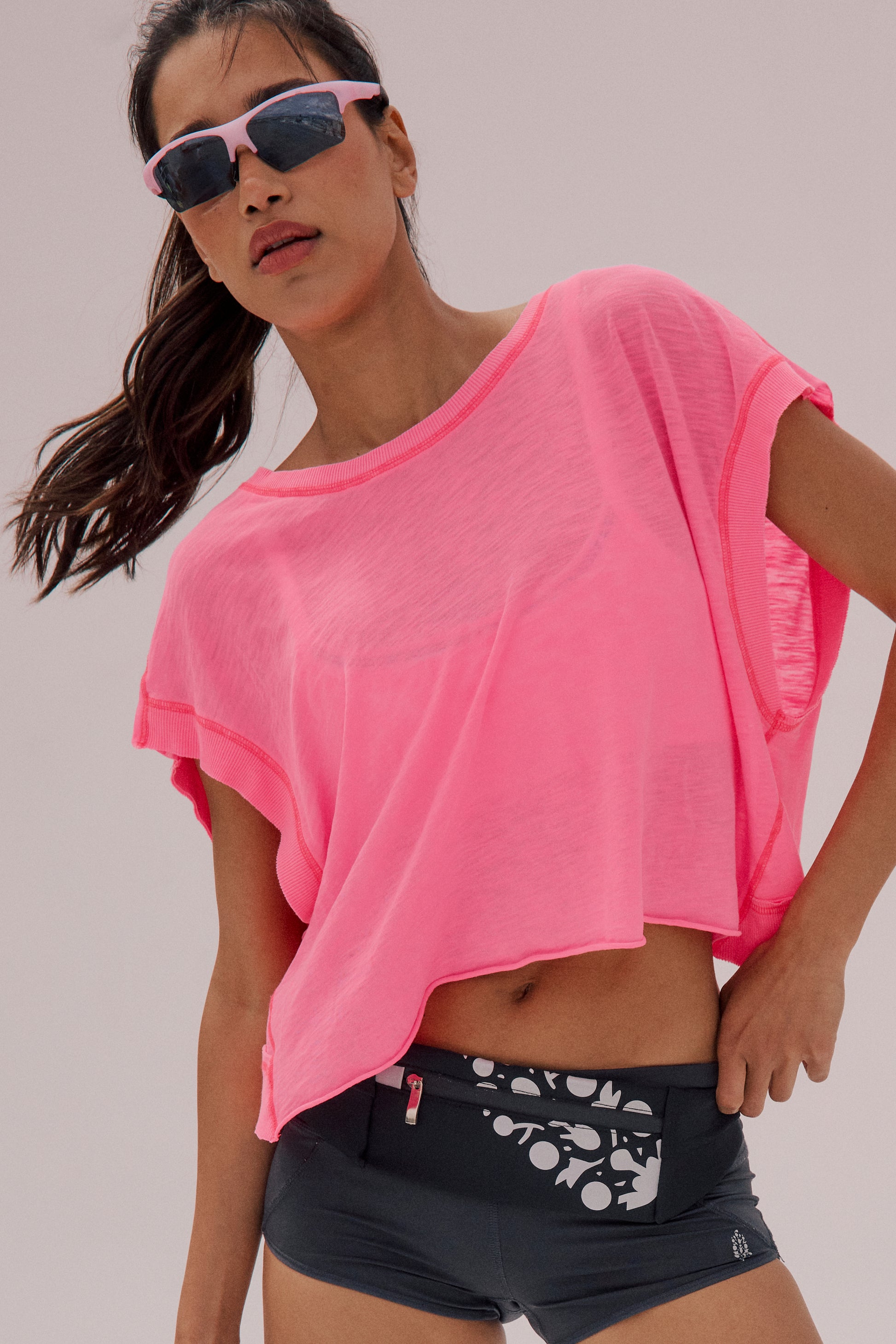 A woman confidently poses against a light background wearing pink sunglasses, black athletic shorts with a white abstract pattern, and the FP Movement My Time Tee in pink.