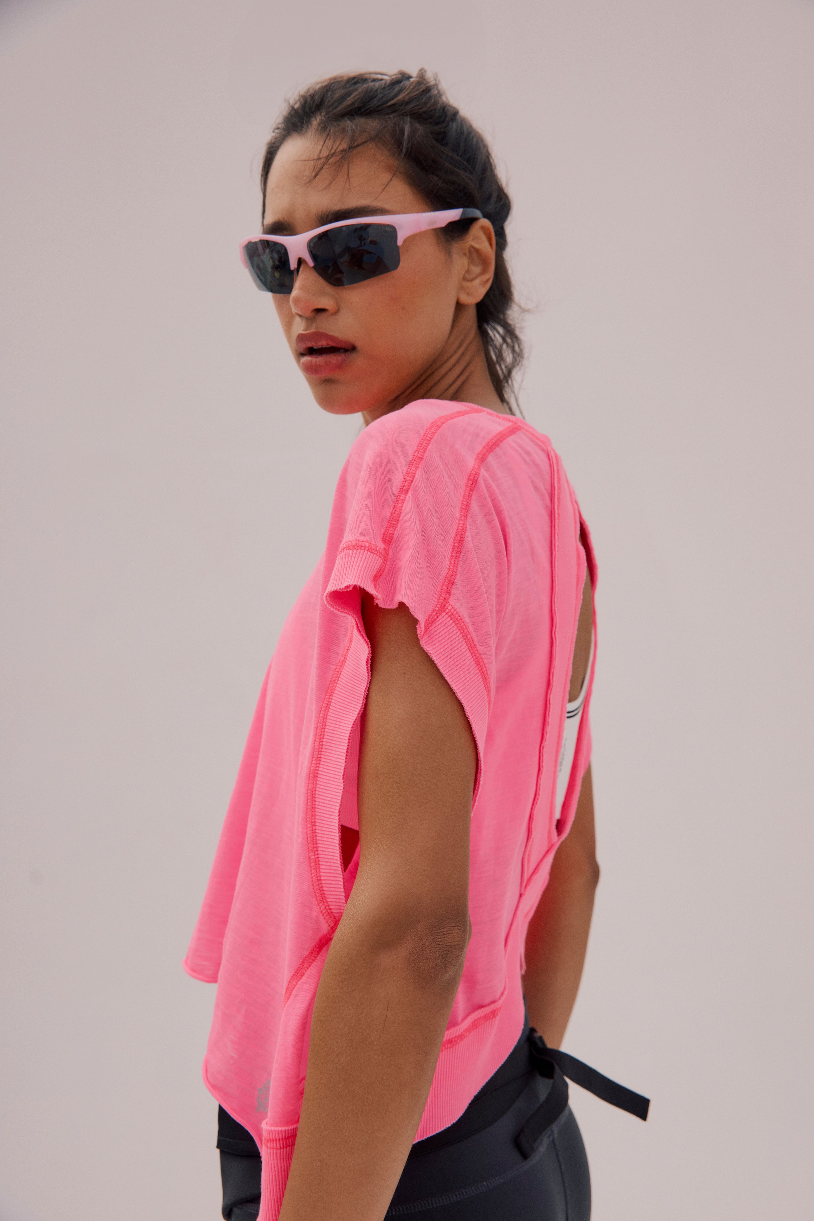 A person wearing the FP Movement My Time Tee—a bright pink, loose-fit top with dropped sleeves and a wide neckline—plus black sunglasses and leggings stands against a plain light background, glancing over their shoulder at the camera.