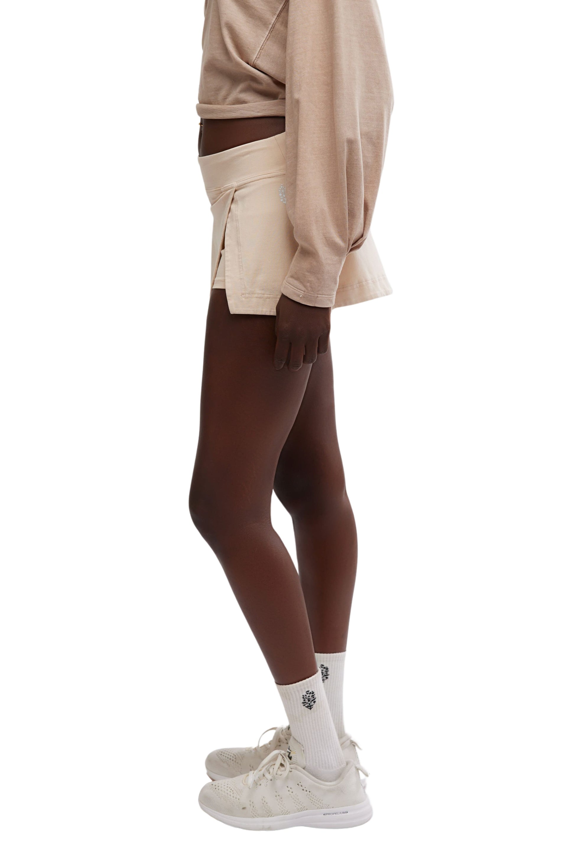 A person stands in profile wearing a beige long-sleeve shirt, the FP Movement Hot Shot Mini Skort in a matching high-rise fit with an elastic waistband, white crew socks with a small black design, and white sneakers against a plain white background.
