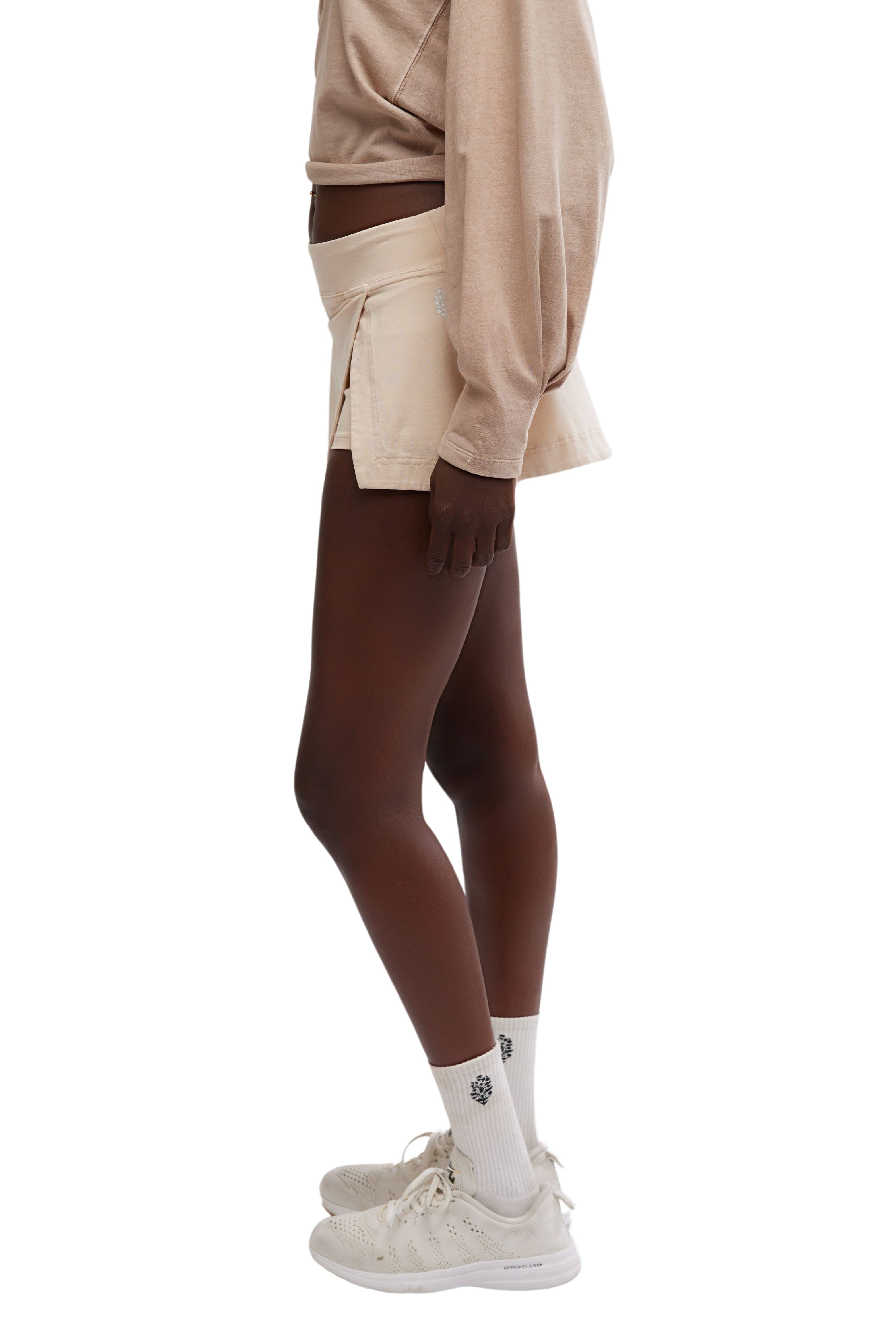A person stands in profile wearing a beige long-sleeve shirt, the FP Movement Hot Shot Mini Skort in a matching high-rise fit with an elastic waistband, white crew socks with a small black design, and white sneakers against a plain white background.