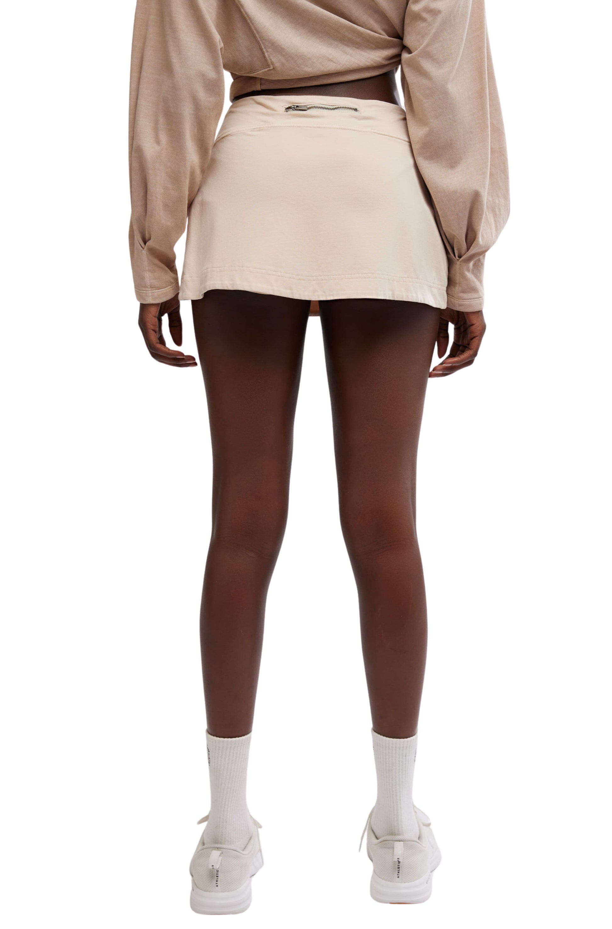 A person stands with their back to the camera, wearing FP Movement's Hot Shot Mini Skort and a beige cotton long-sleeve top, paired with white socks and shoes.