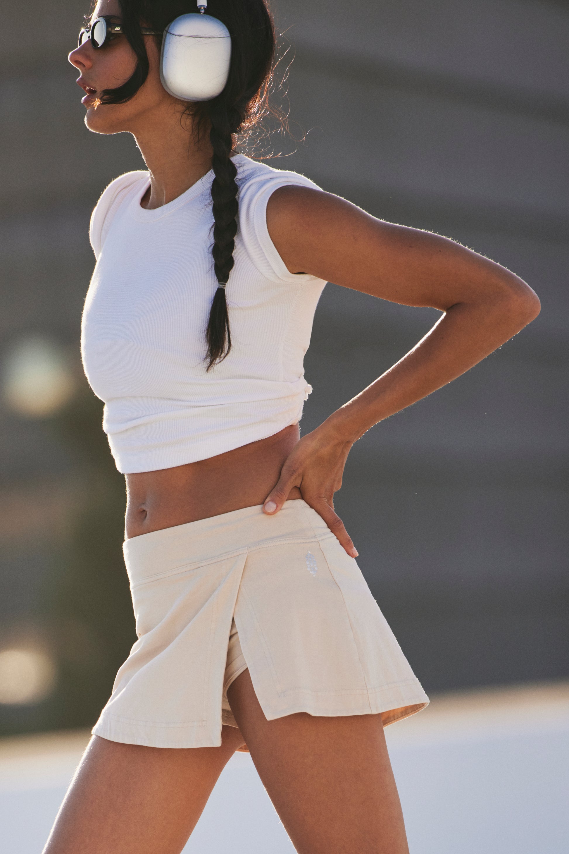 A woman with a braid wears large headphones, a white crop top, and the FP Movement Hot Shot Mini Skort while standing outdoors with one hand on her hip.