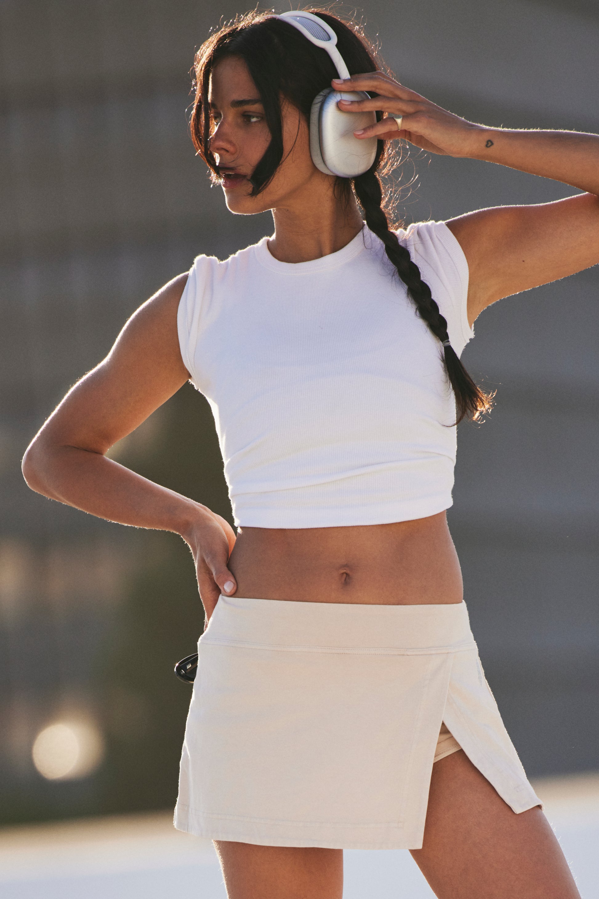 A woman outdoors wears large over-ear headphones, a white crop top, and the FP Movement Hot Shot Mini Skort. She has a braid, one hand on her hip and the other adjusting her headphones.
