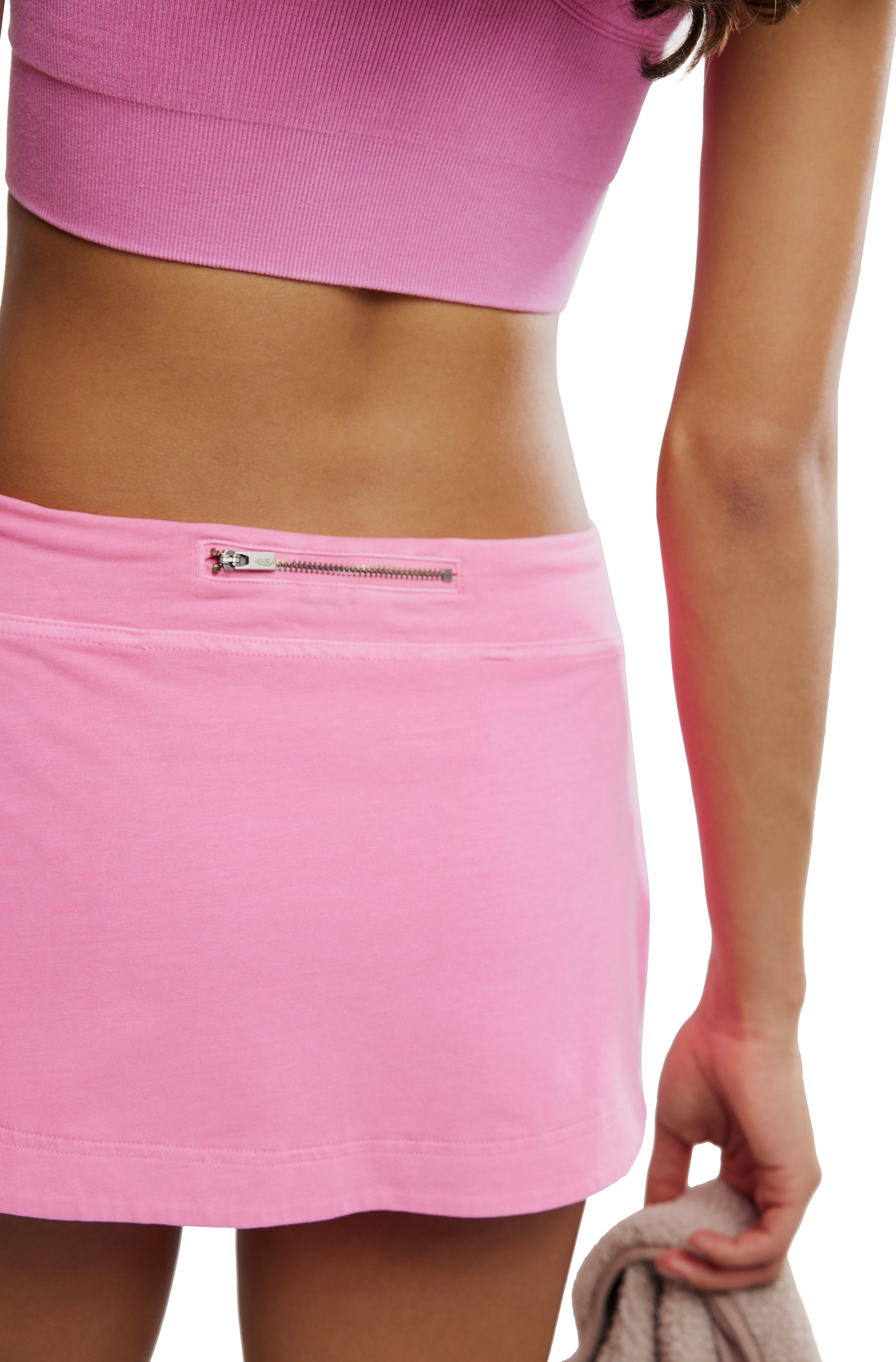 A person wears the FP Movement Hot Shot Mini Skort, a pink high-rise piece with a small zipper pocket on the waistband, paired with a pink crop top and holds a beige garment in their right hand. Their midsection and arm are visible.