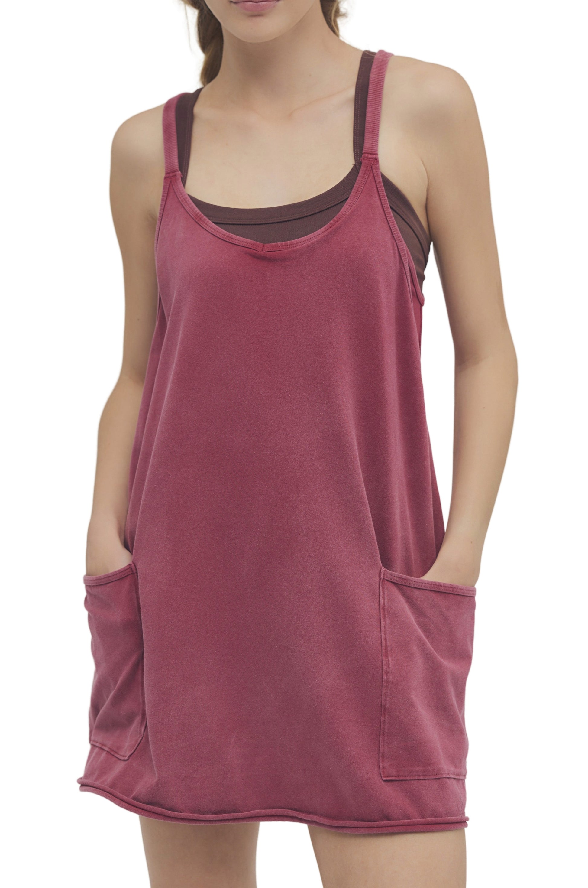 A woman wears the FP Movement Hot Shot Mini, a sleeveless rose mini dress with two large front pockets and a scoop neckline, styled over a brown tank top. The casual, loose fit is shown in an image cropped at the neck and above the knees.