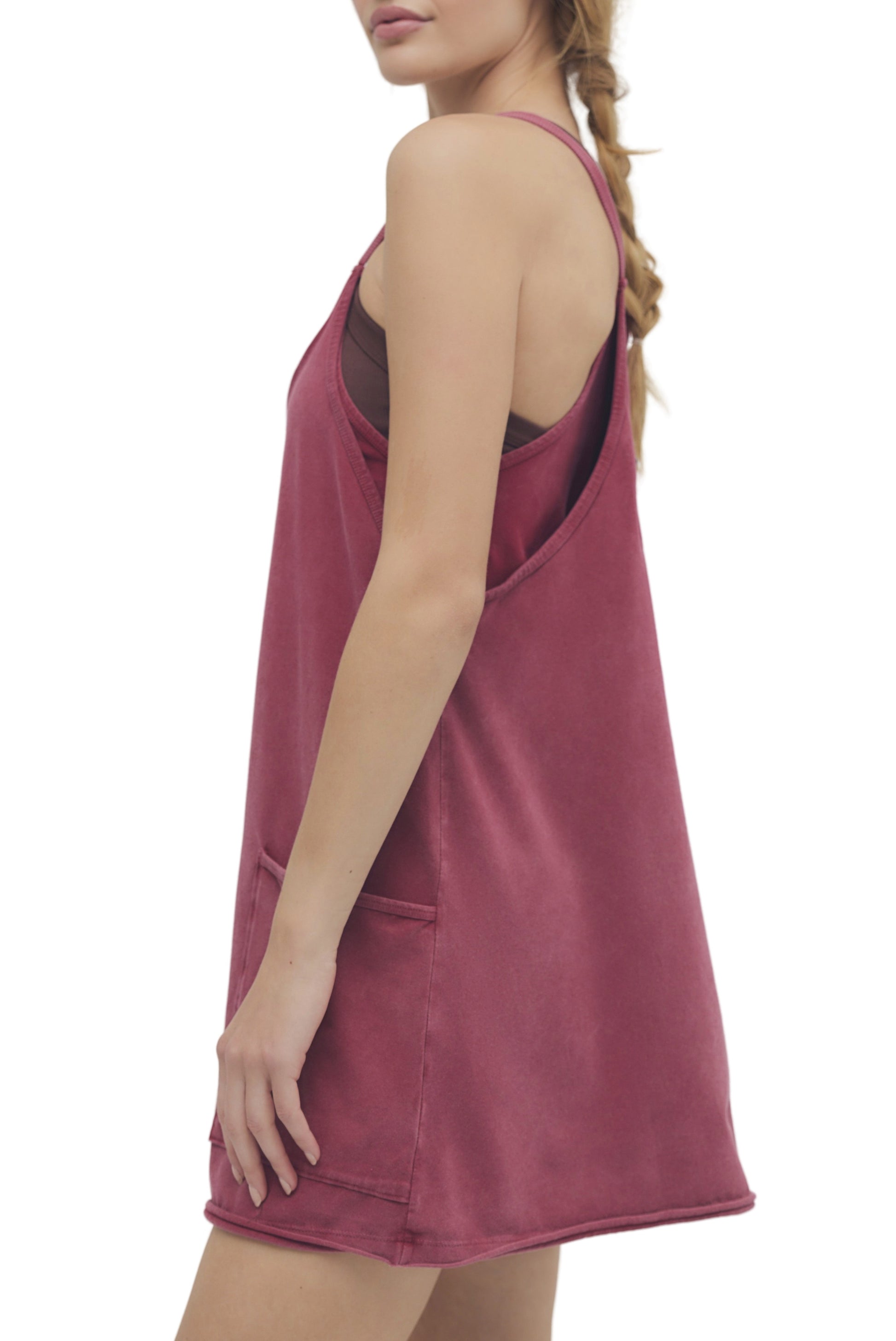 A person with a braid is shown in profile wearing the FP Movement Hot Shot Mini, a loose-fitting, sleeveless dark pink dress with pockets and a scoop neckline over a brown top. The background is white.