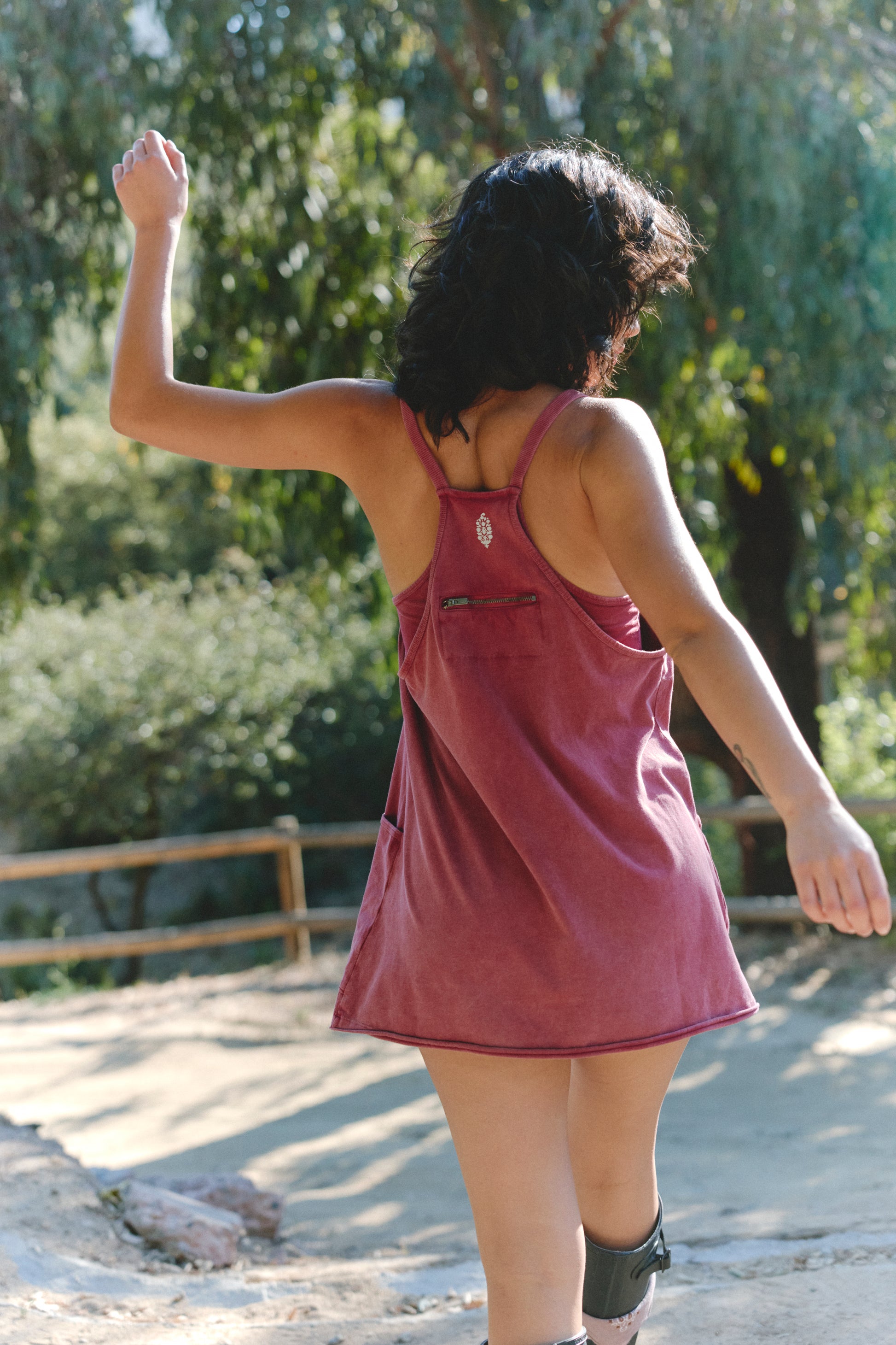 A person with dark hair wears the FP Movement Hot Shot Mini, a sleeveless pink scoop-neck mini dress, outdoors on a sunny day with one arm raised. Trees and a wooden fence can be seen in the background.