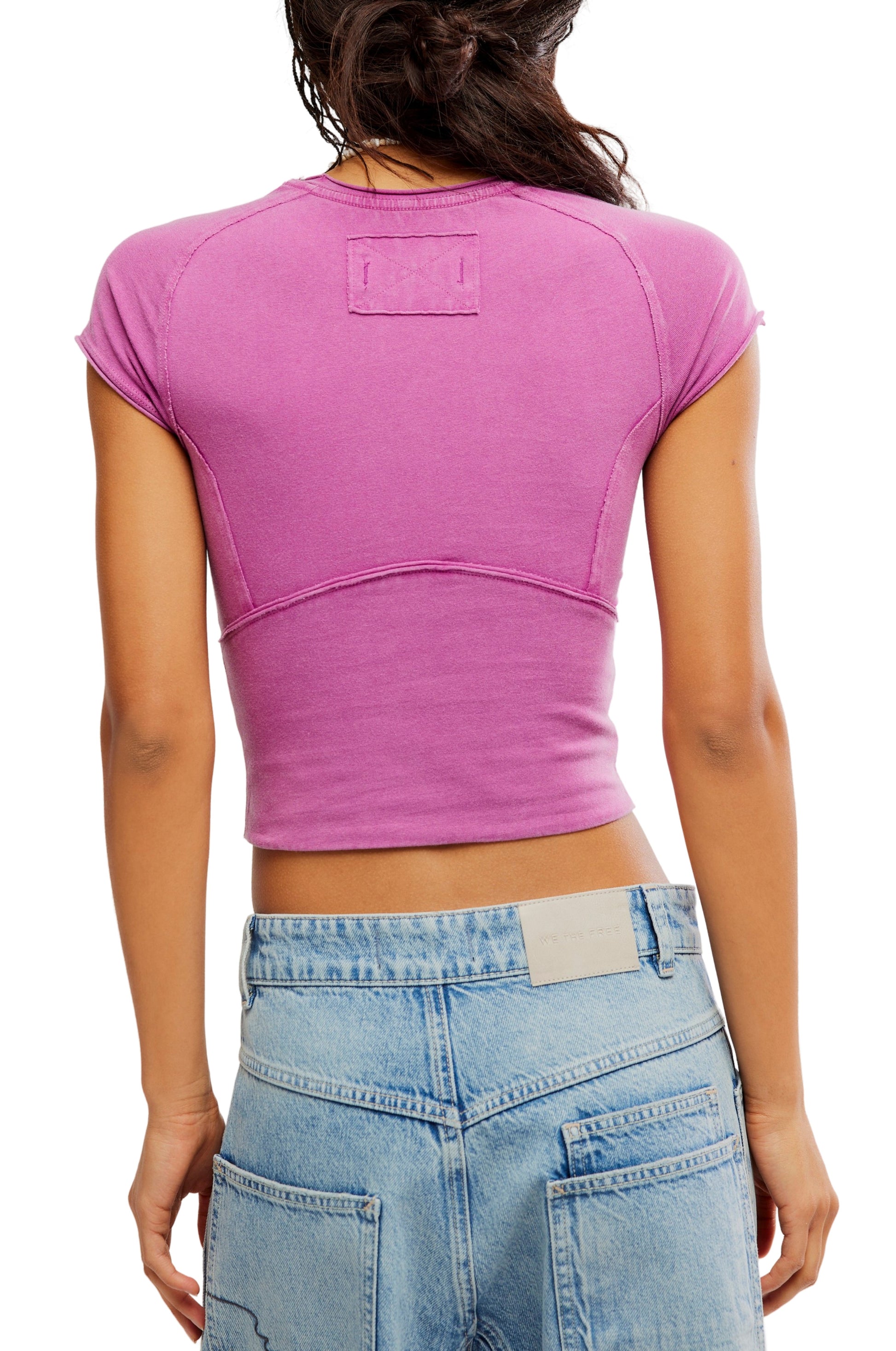 A person shown from the back wears the Free People Protagonist Tee, a vintage-inspired fitted crop top, paired with high-waisted blue jeans against a plain white background.
