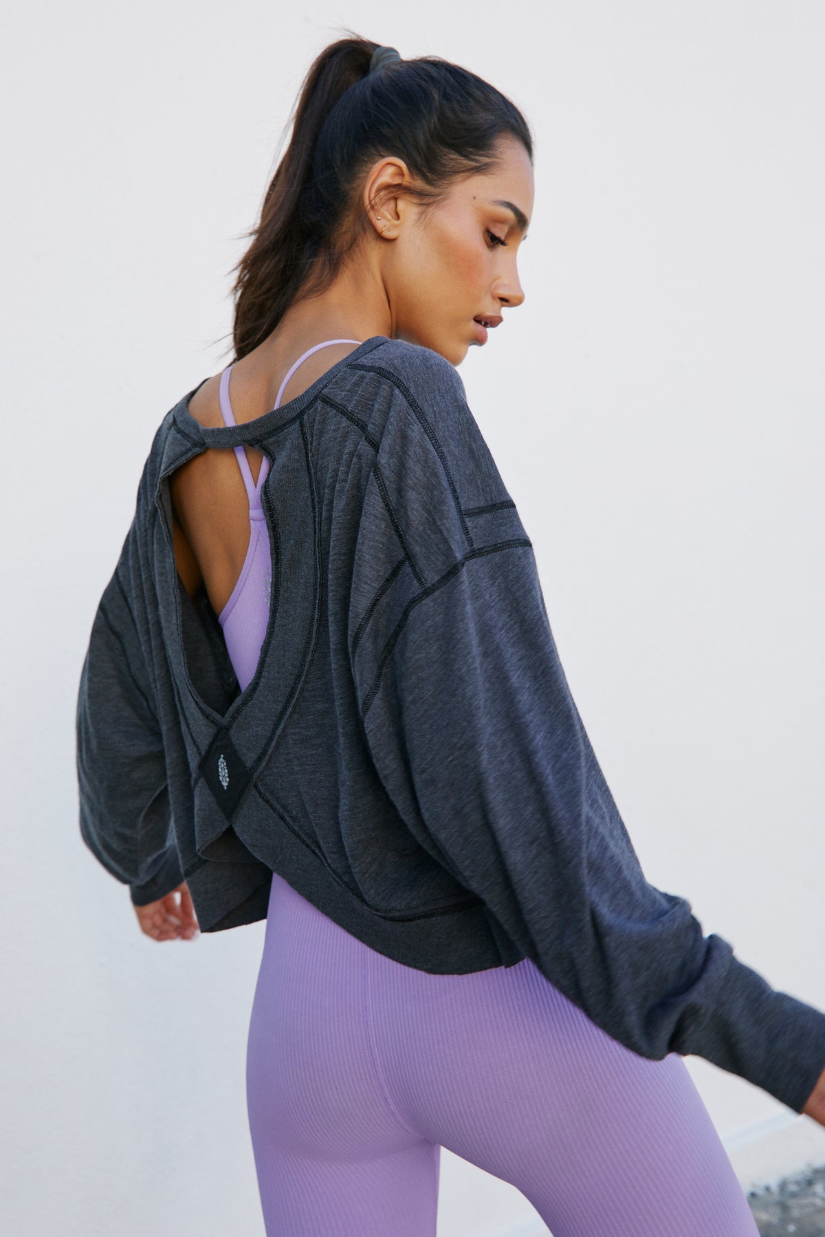 Woman wearing black layered open-back jacket over lavender ribbed leggings, shown from side view.