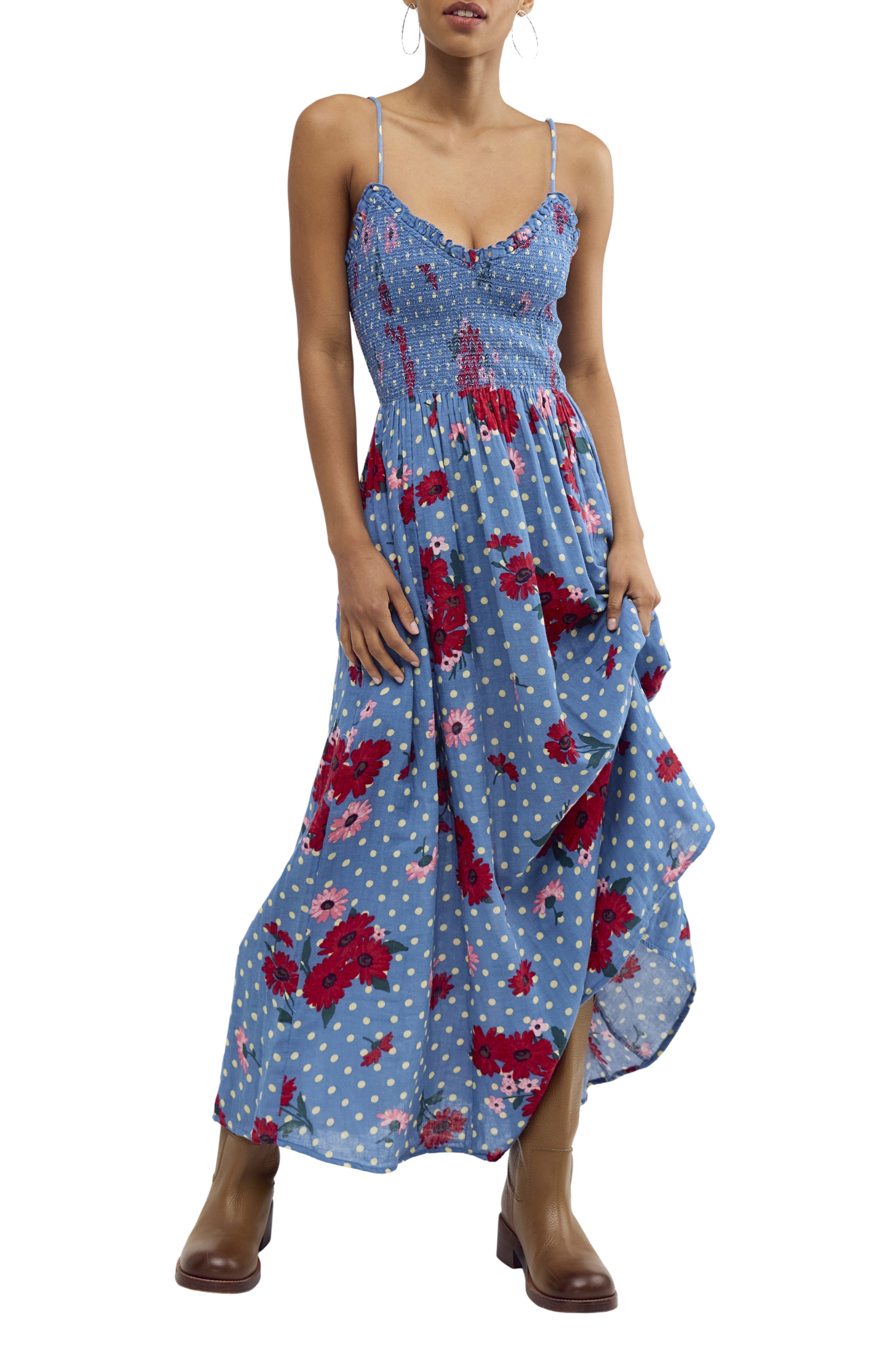 A woman wears the Free People Sweet Nothings Midi in Blue Multi, a sleeveless dress with red floral and white dot patterns, a smocked bodice, brown ankle boots, and hoop earrings, holding the skirt with one hand.