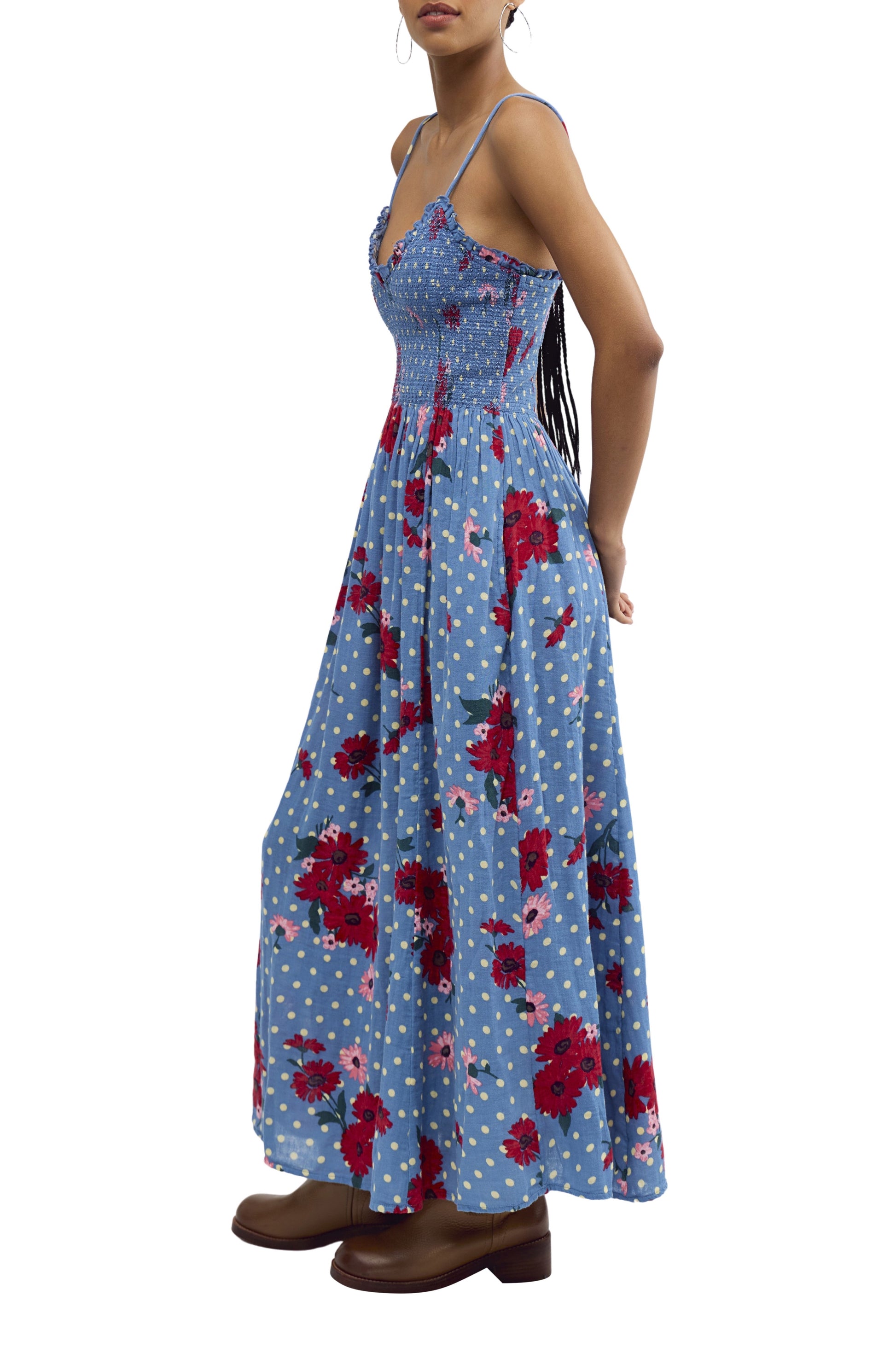 A woman with long hair wears the Free People Sweet Nothings Midi, a blue sleeveless dress with a smocked bodice, red floral and white polka dot patterns, paired with brown boots. She stands sideways, arms relaxed.