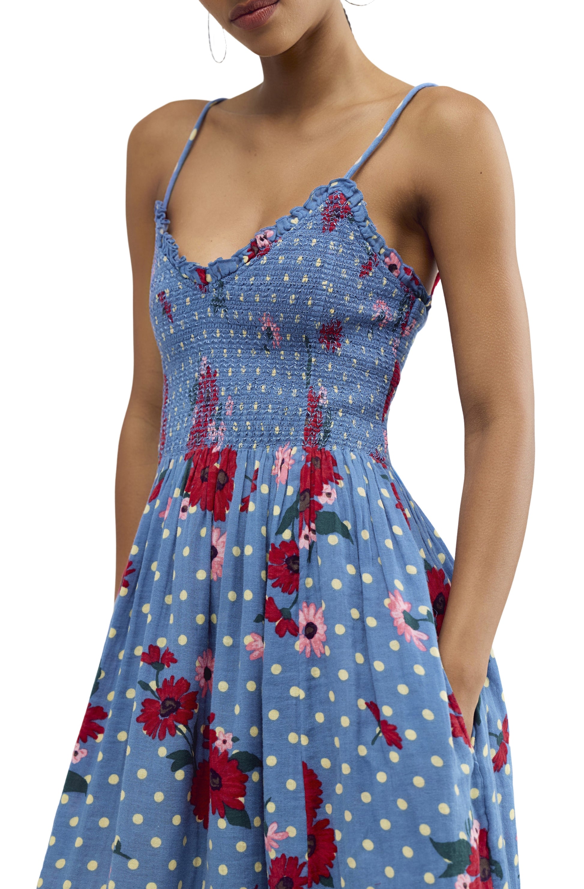 A woman wears the Sweet Nothings Midi by Free People—a sleeveless, light blue cotton dress with yellow polka dots, red floral prints, thin straps, smocked bodice, and side pockets.