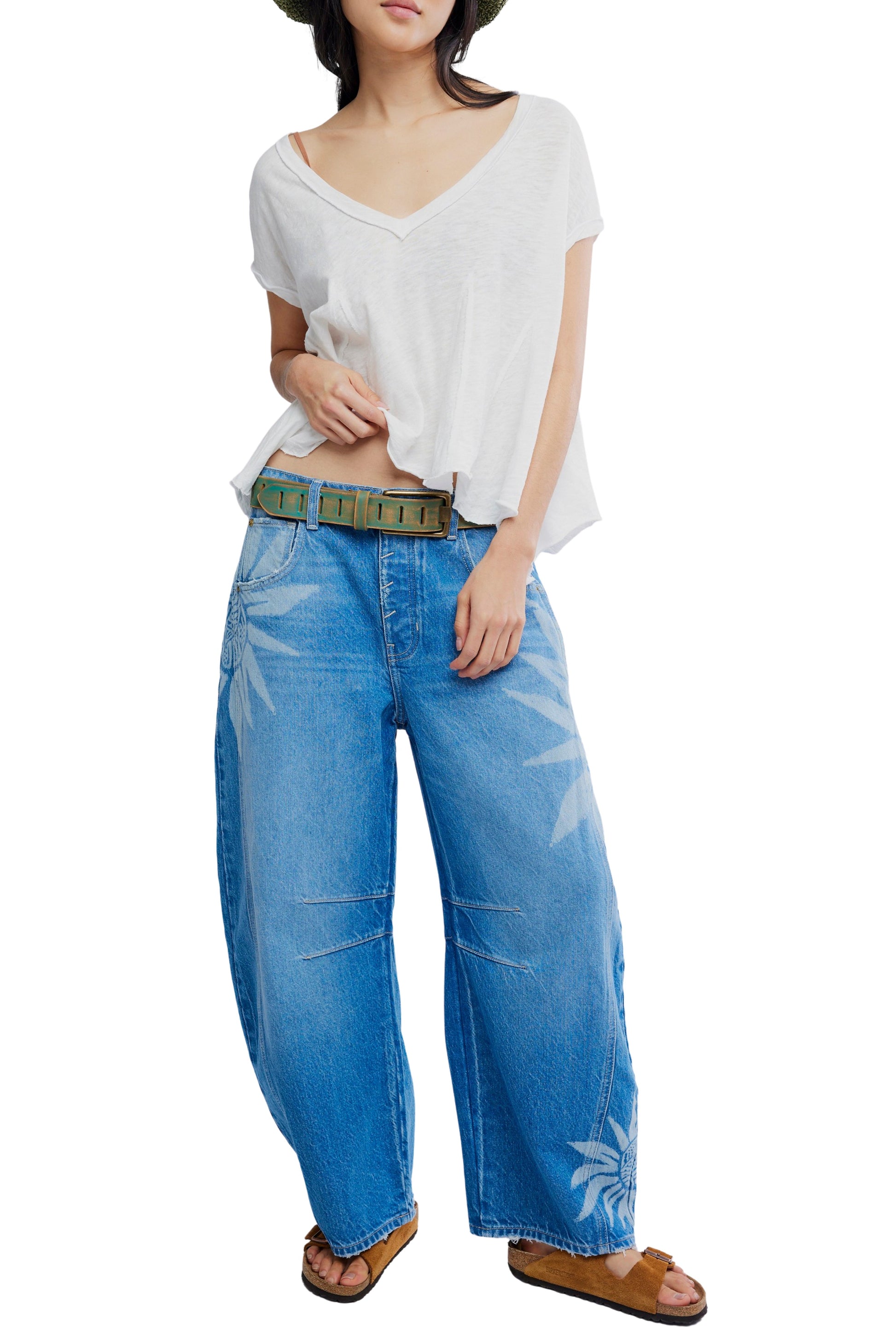 A woman wears a loose white V-neck T-shirt, a green belt, brown sandals, a dark hat, and Free People’s Good Luck Printed Barrel Jean in Medium Denim with one hand in her pocket and the other adjusting her shirt.