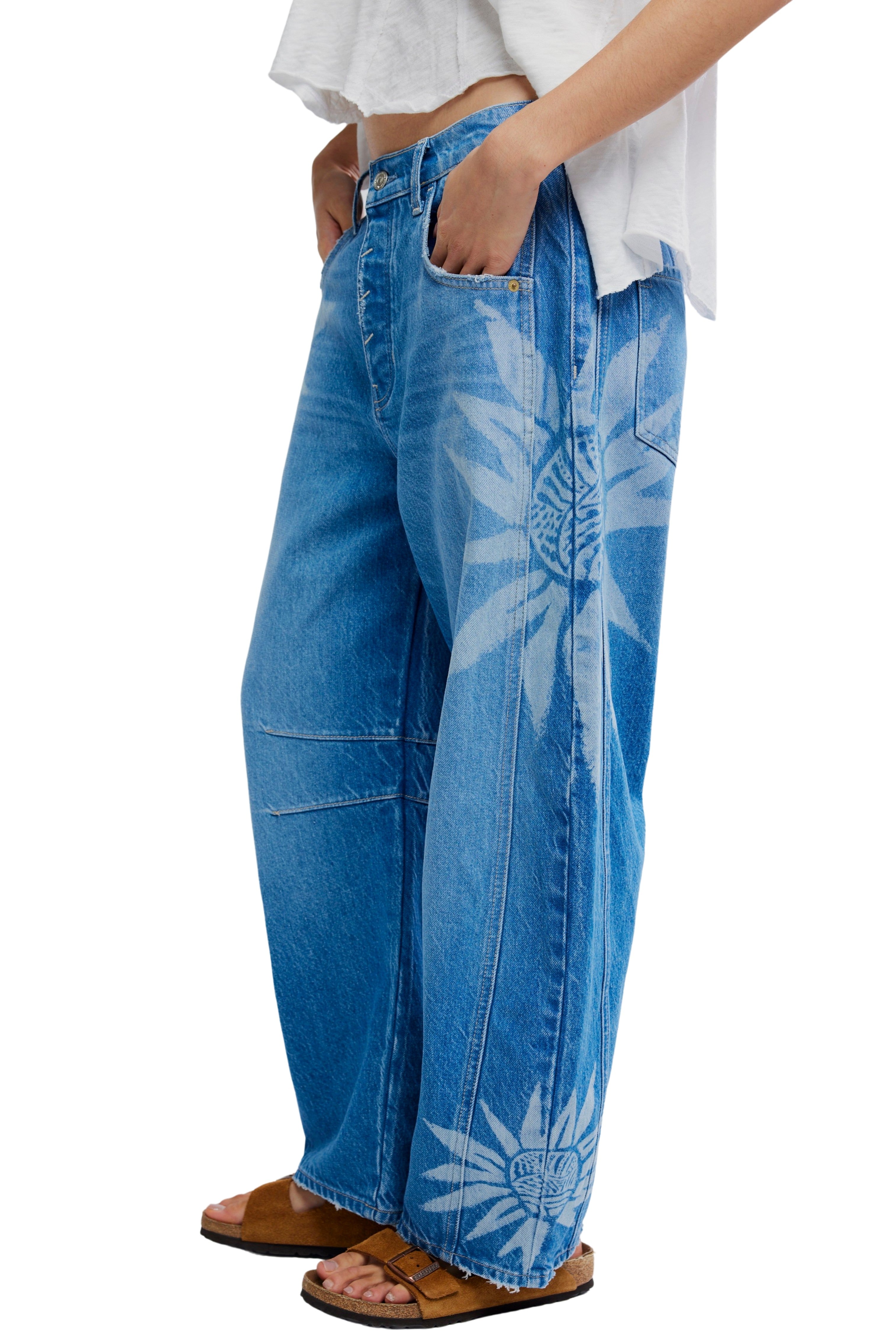 A person wears Free People’s Good Luck Printed Barrel Jean—relaxed-fit, wide-leg blue jeans with bold white floral prints in recycled cotton denim—paired with a loose white top and brown sandals. The image is cropped at the shoulders.