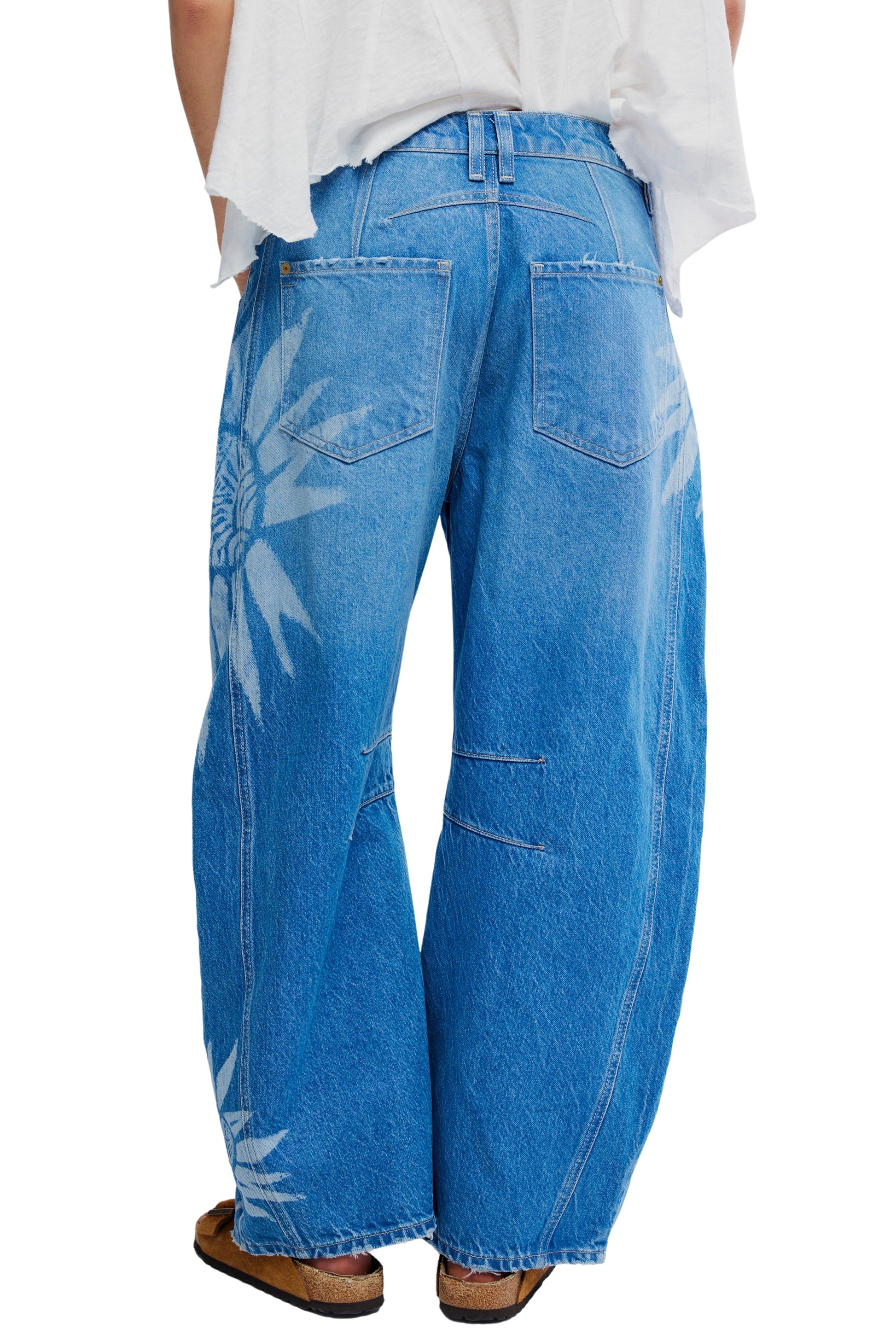A person models the Free People Good Luck Printed Barrel Jean, featuring a relaxed fit and white sunburst designs on recycled cotton denim; photographed from the back to showcase the rear details, paired with sandals and a loose white shirt.
