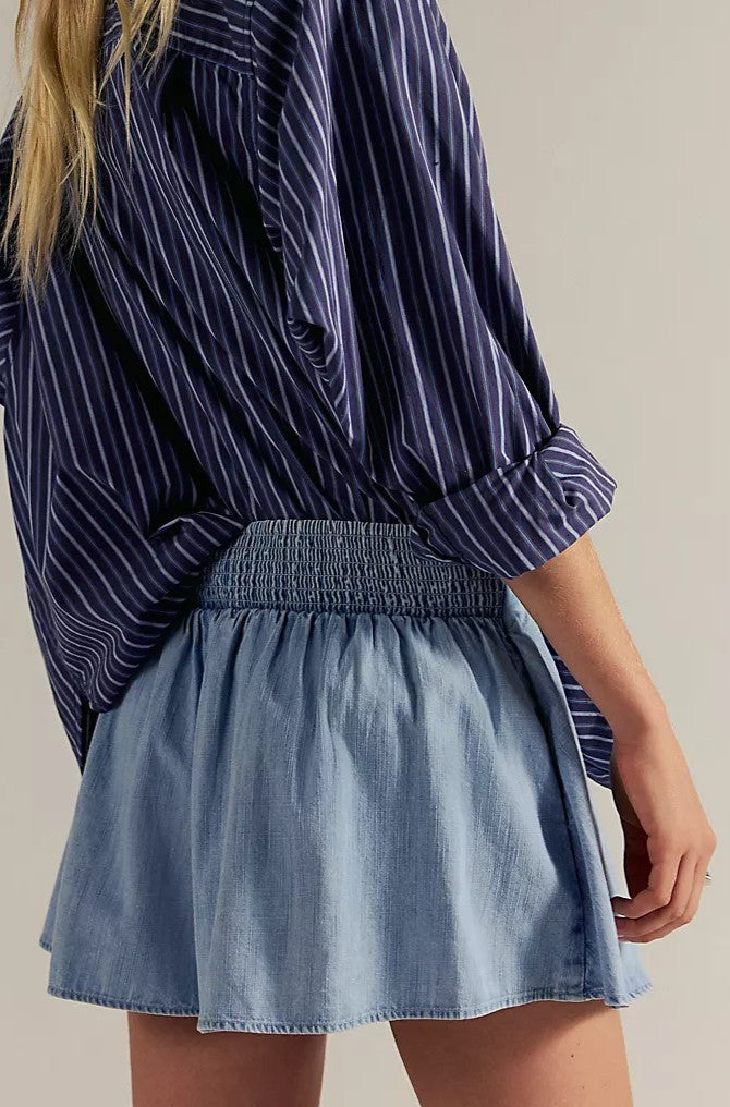 Wearing the Free People Paradise Found Denim Skort—a light blue, 100% cotton, pull-on flared style—the person stands sideways with one arm relaxed by their side, paired with a blue and white striped long-sleeve shirt.