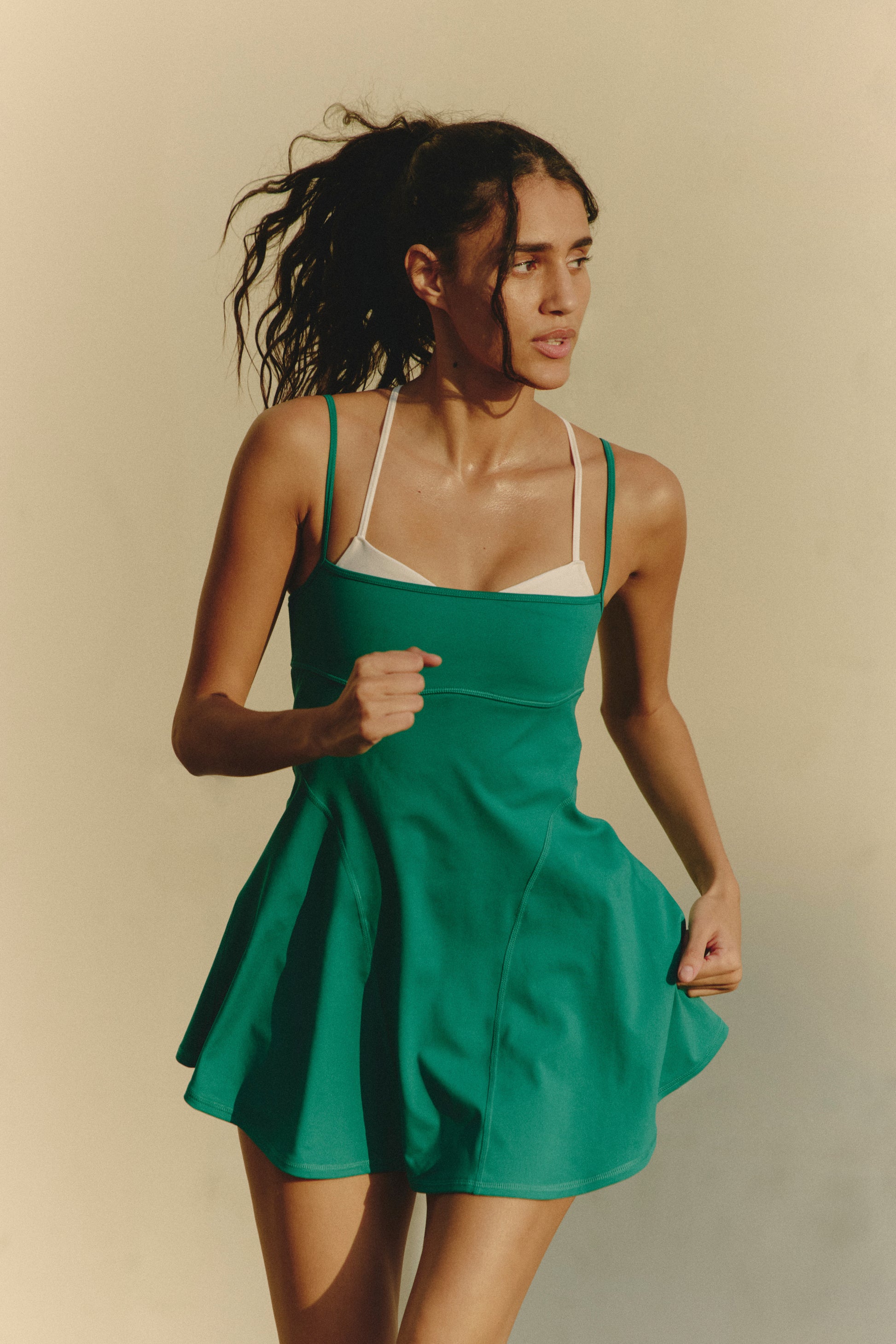 A woman with long dark hair in a ponytail runs outdoors, wearing the FP Movement Baseline Tennis Dress—a green, thin-strap A-line dress layered over a white sports bra—against a plain light beige background.