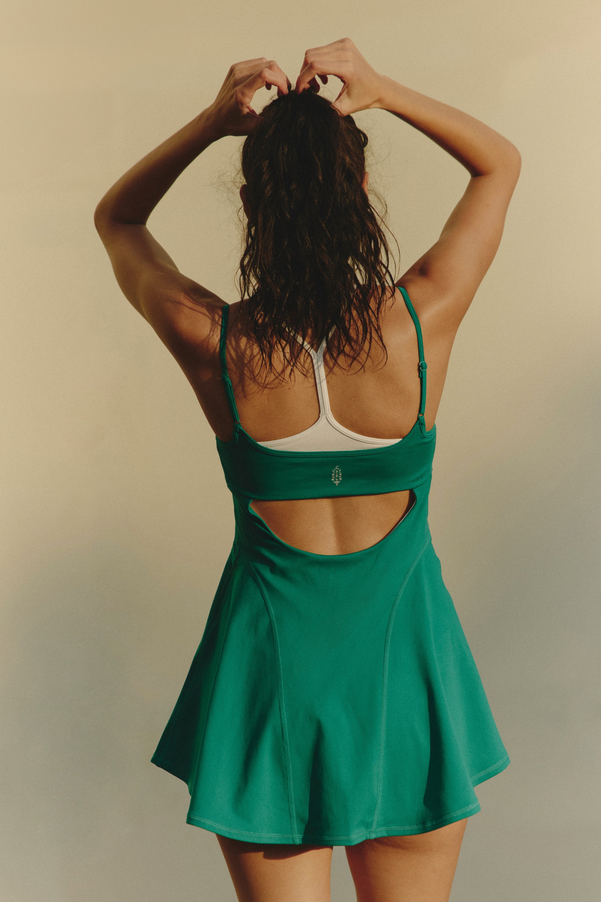 A woman faces away, tying her hair up. She wears the green FP Movement Baseline Tennis Dress with built-in shorts and a white sports bra underneath, set against a plain neutral backdrop.