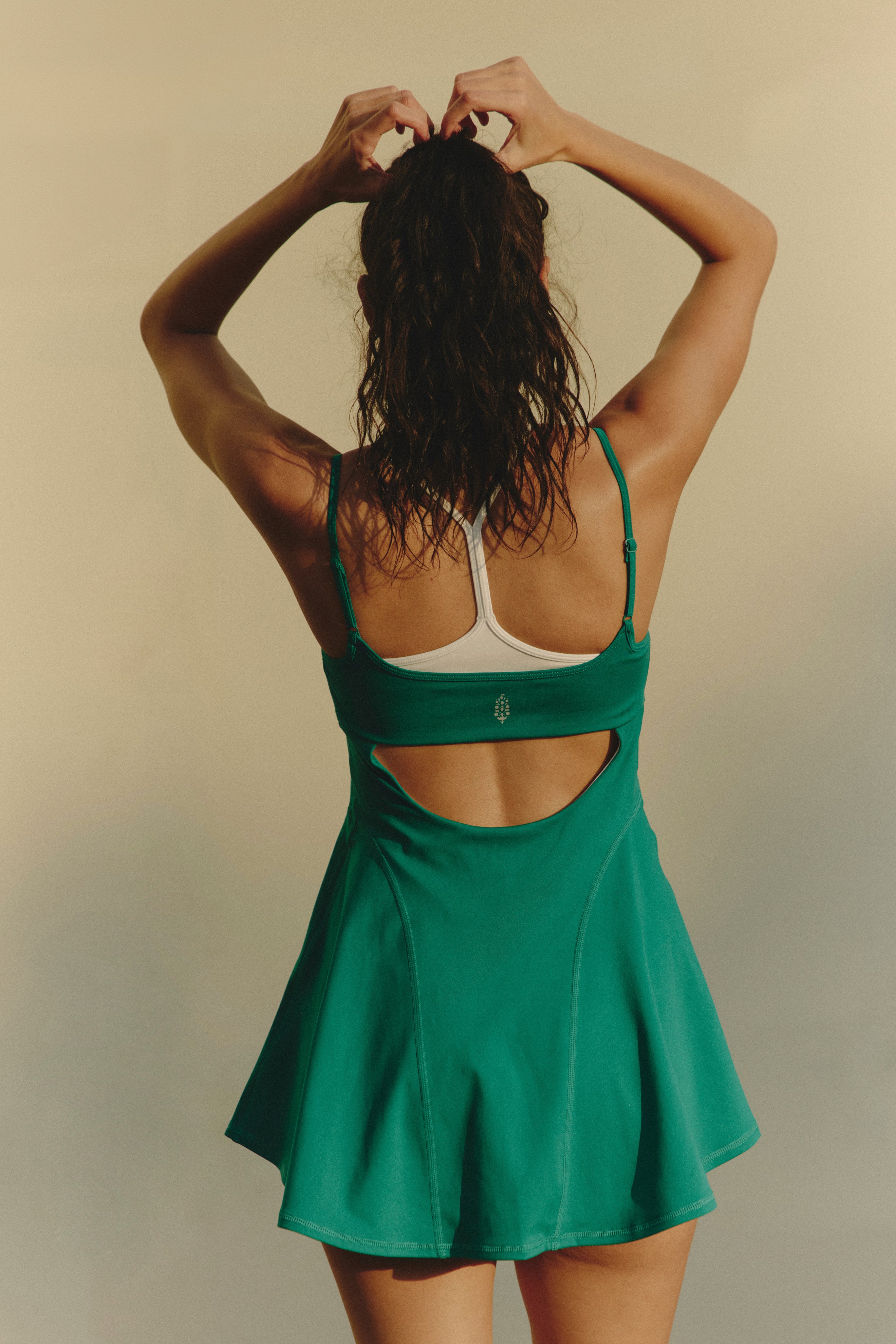 A woman faces away, tying her hair up. She wears the green FP Movement Baseline Tennis Dress with built-in shorts and a white sports bra underneath, set against a plain neutral backdrop.