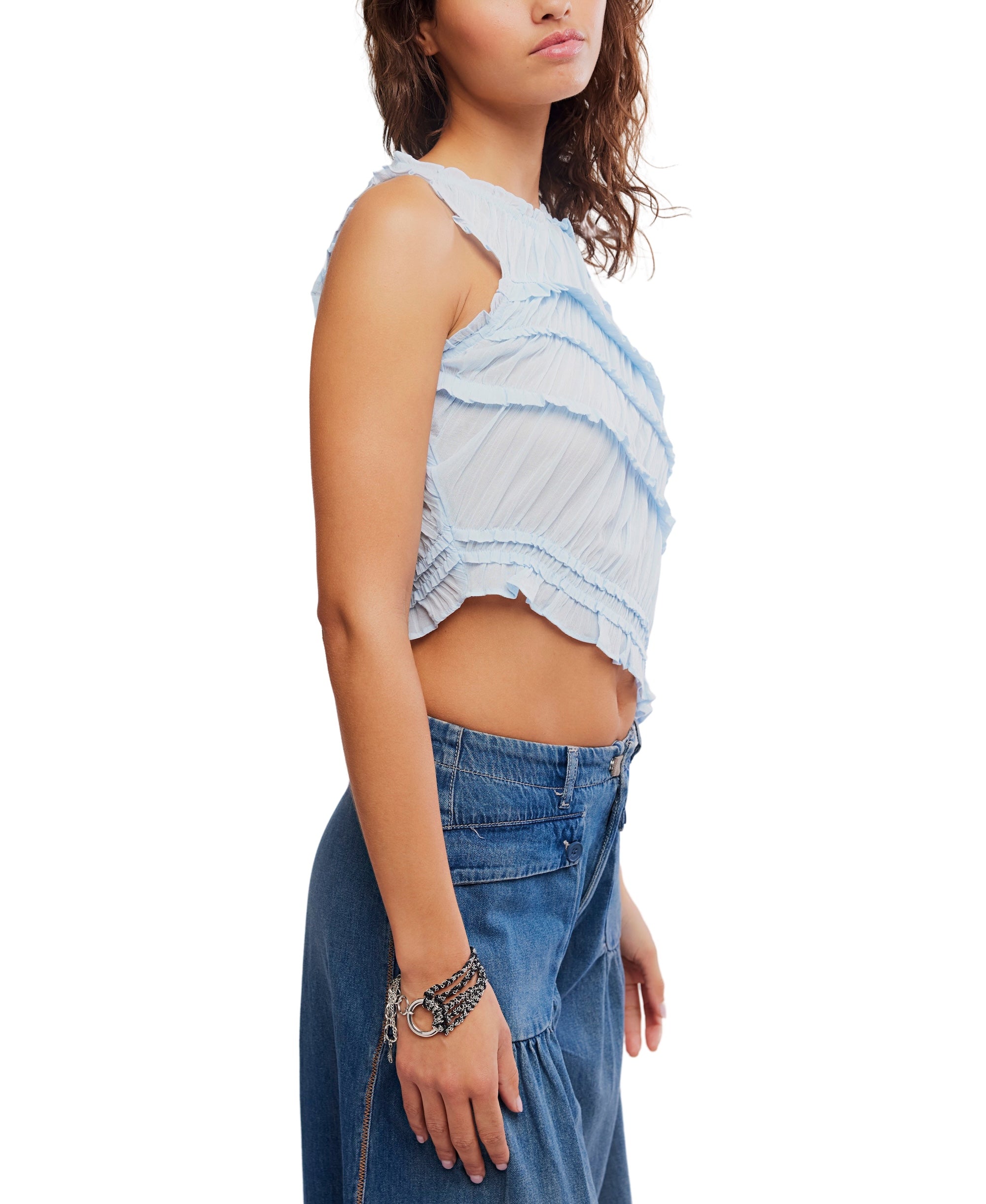 A woman in the Free People Pink Sands Smocked Top stands in profile against a white background, wearing a sleeveless, ruffled crop top with a jewel neck and high-waisted blue jeans, accessorized with a silver bracelet.