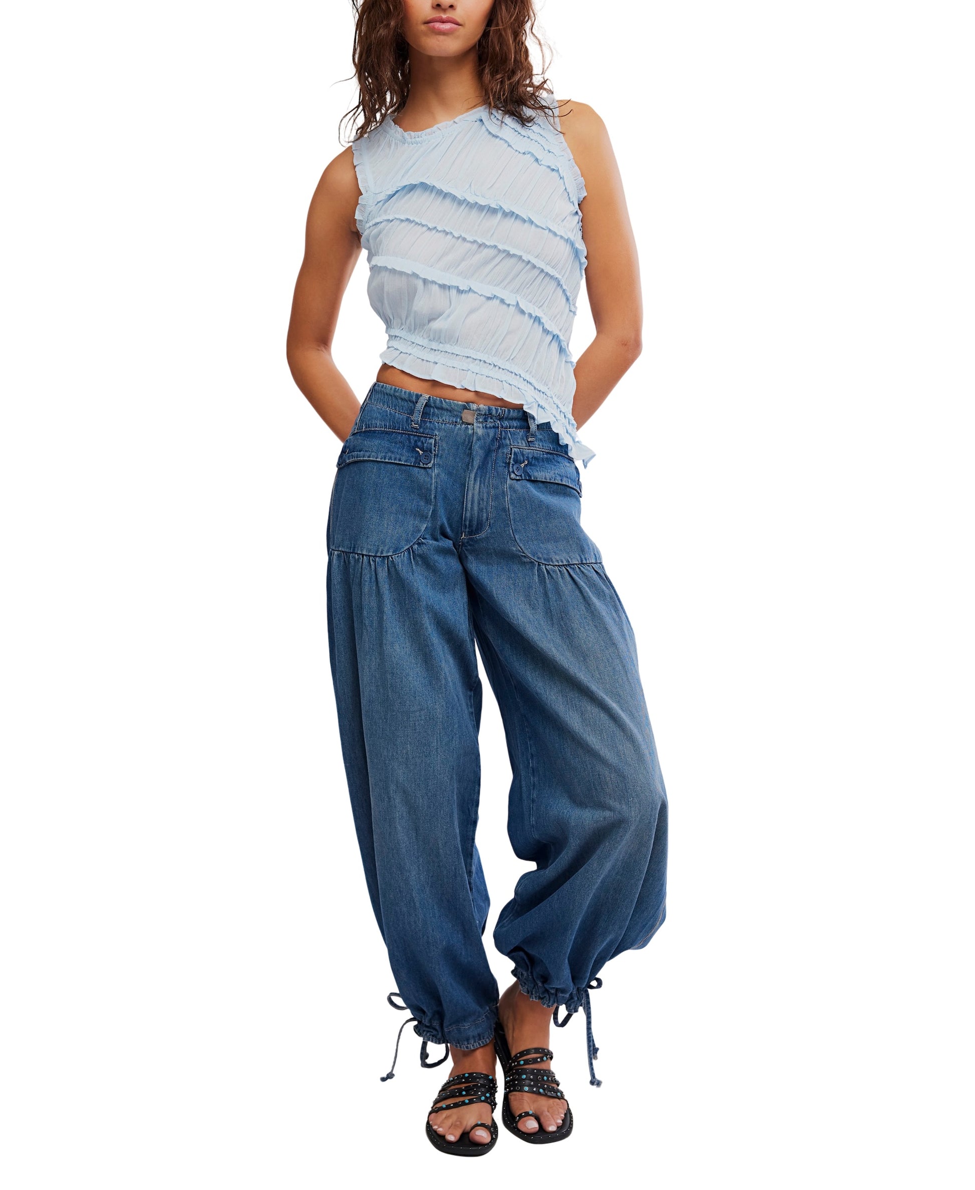 A woman wears the Free People Pink Sands Smocked Top, a sleeveless light blue viscose-cotton top with ruffles and a jewel neck, paired with loose-fit blue jeans with drawstring hems and black sandals, standing against a white background.