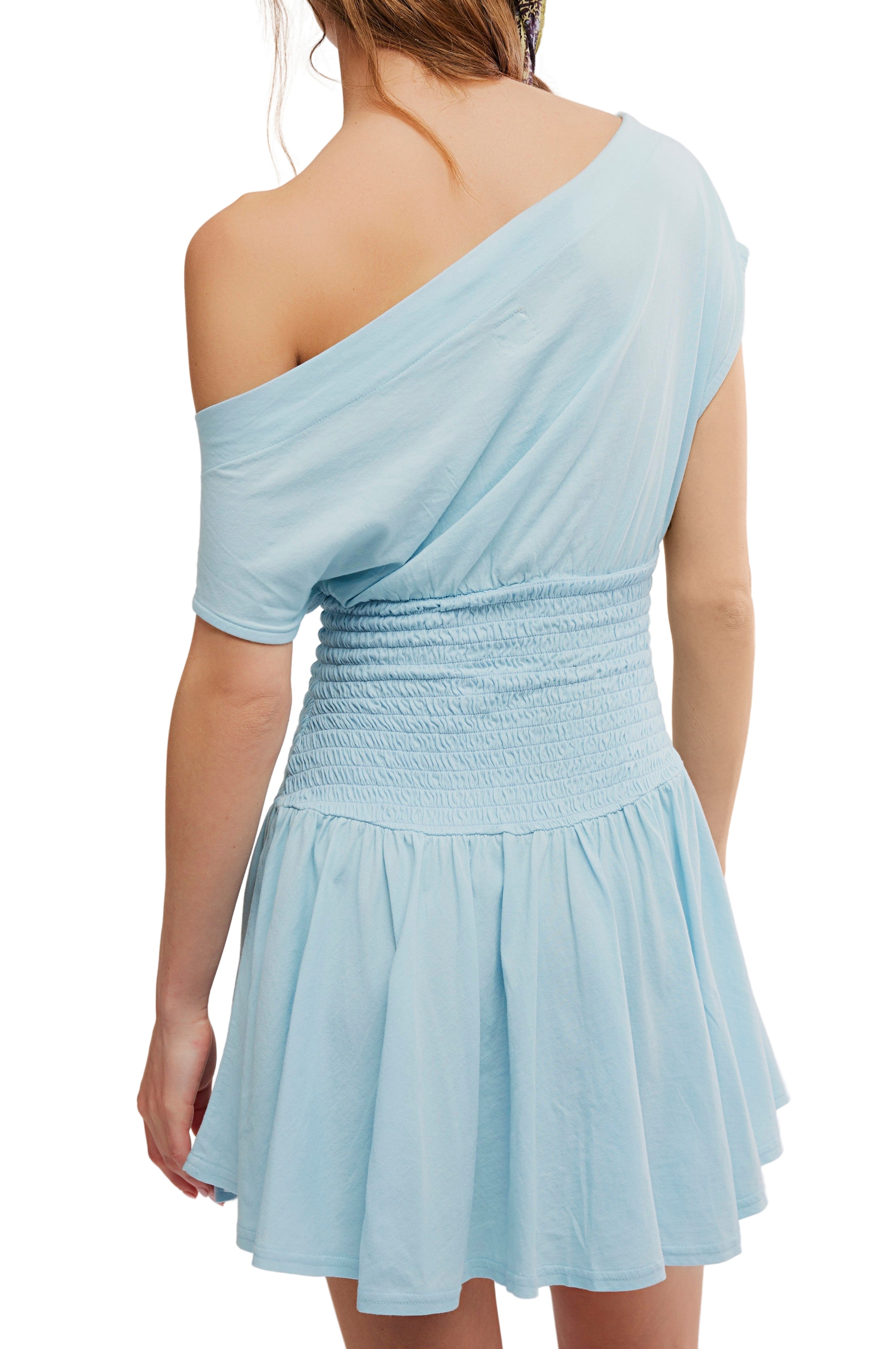 A woman wearing the Free People Scorching Hot Mini—a light blue, off-the-shoulder dress with a smocked bodice and flared skirt, crafted from soft cotton jersey—shown from the back.