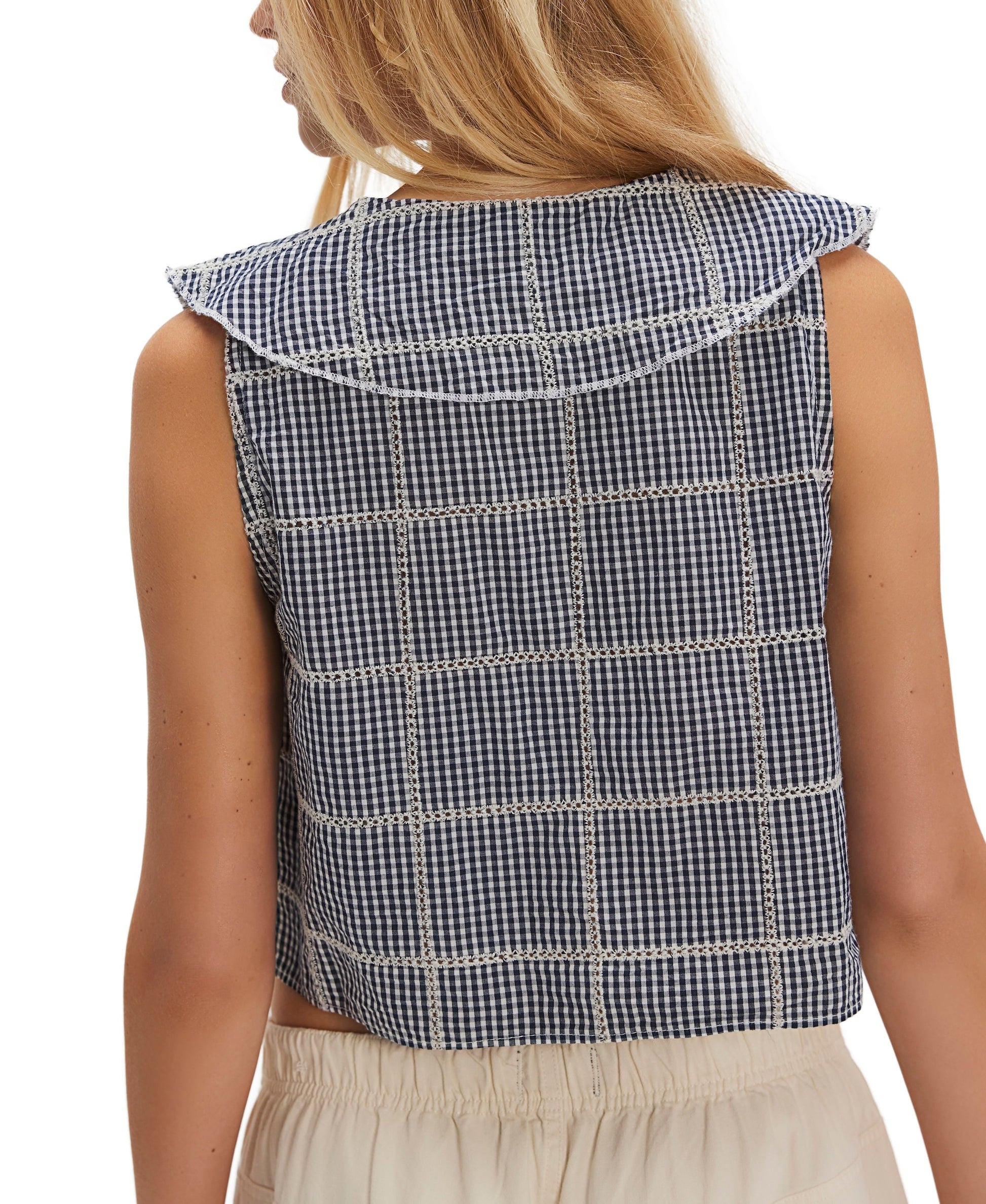 A woman with long blonde hair is seen from behind wearing the Free People Waiting For Me Top, a navy blue and white checkered sleeveless top with a sailor collar, paired with cream-colored pants.