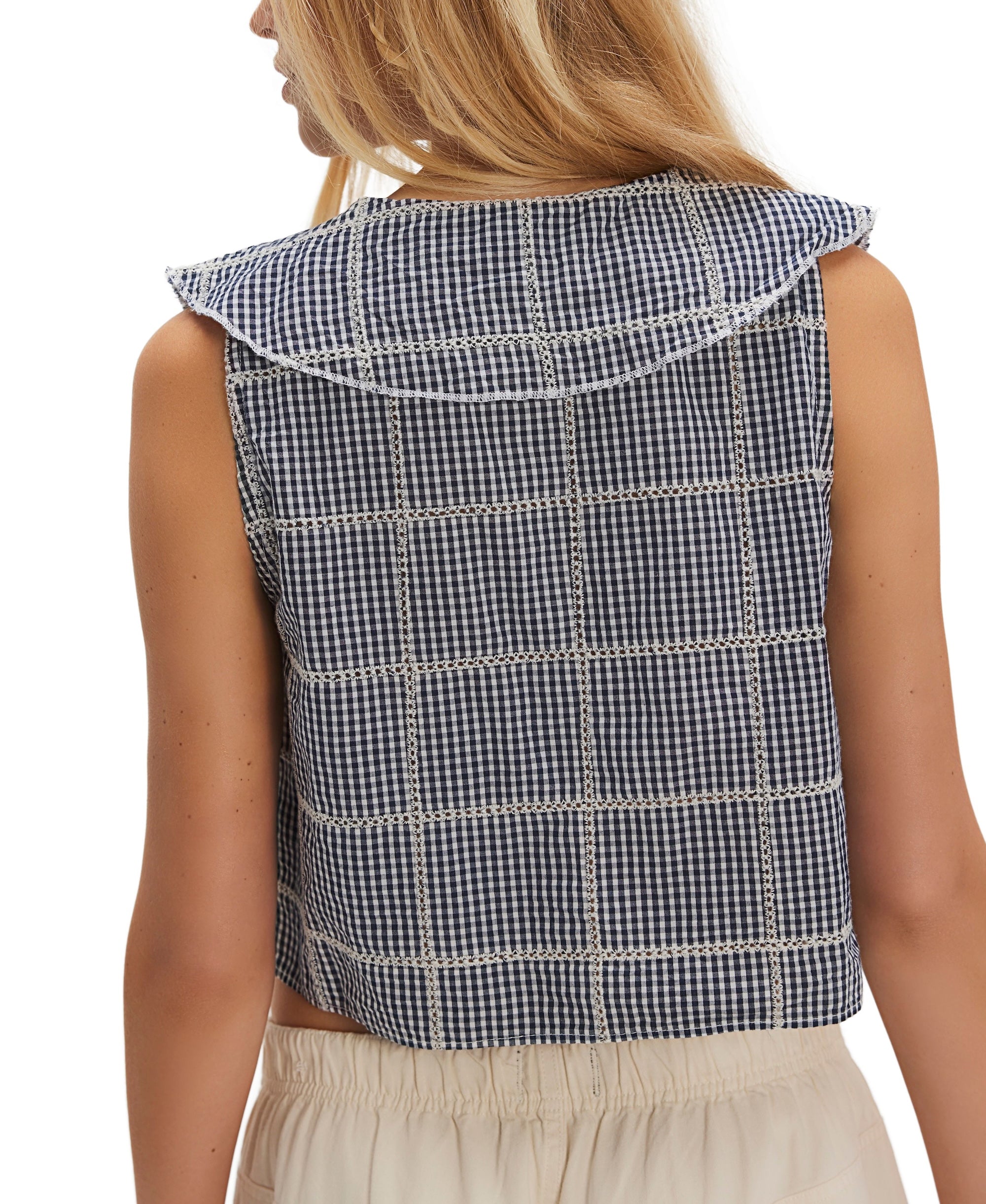 A woman with long blonde hair is seen from behind wearing the Free People Waiting For Me Top, a navy blue and white checkered sleeveless top with a sailor collar, paired with cream-colored pants.