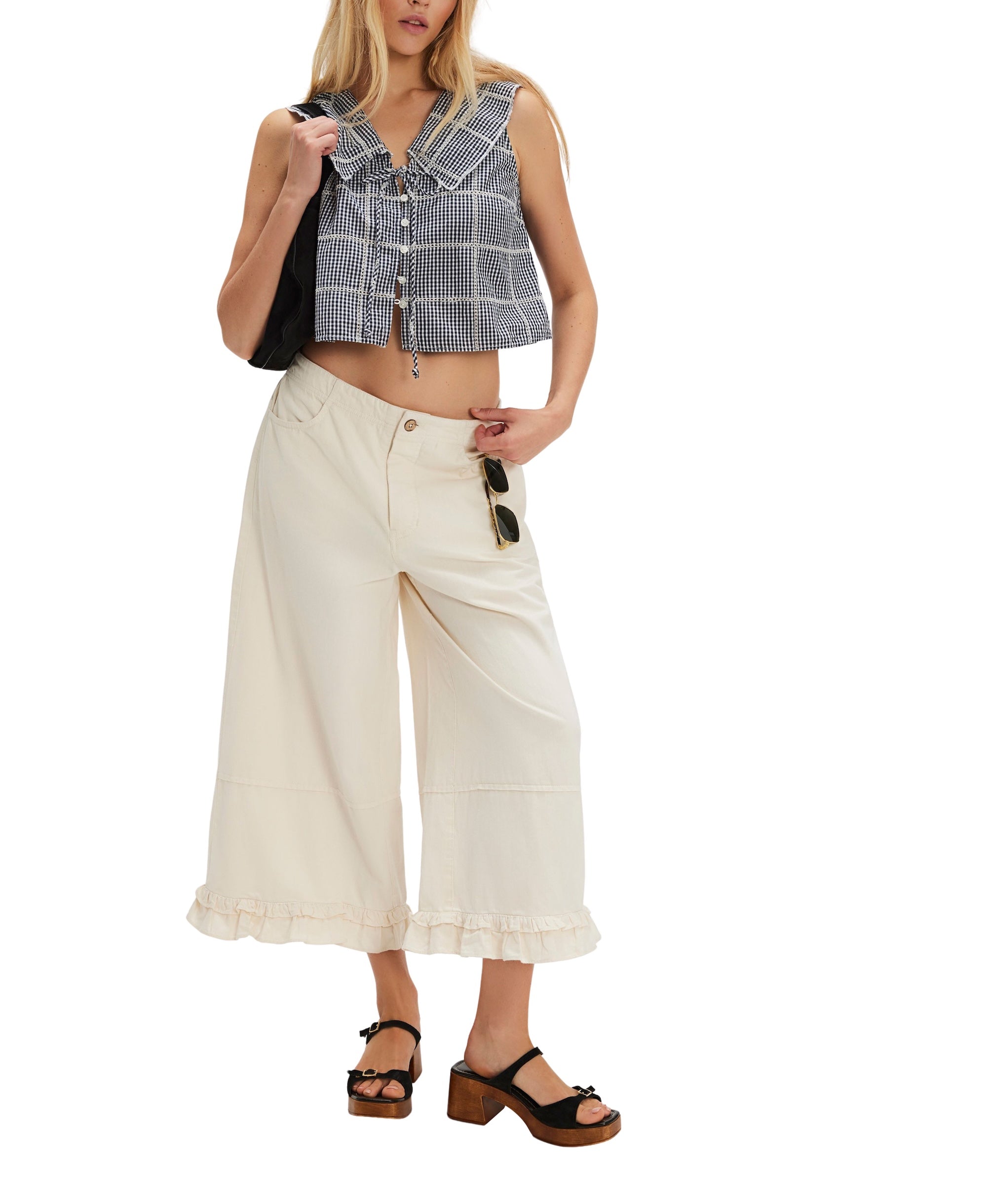 A woman wears the Waiting For Me Top by Free People (a sleeveless black and white checkered crop top), cream wide-leg ruffled-hem pants, black platform sandals, sunglasses, and carries a black shoulder bag.