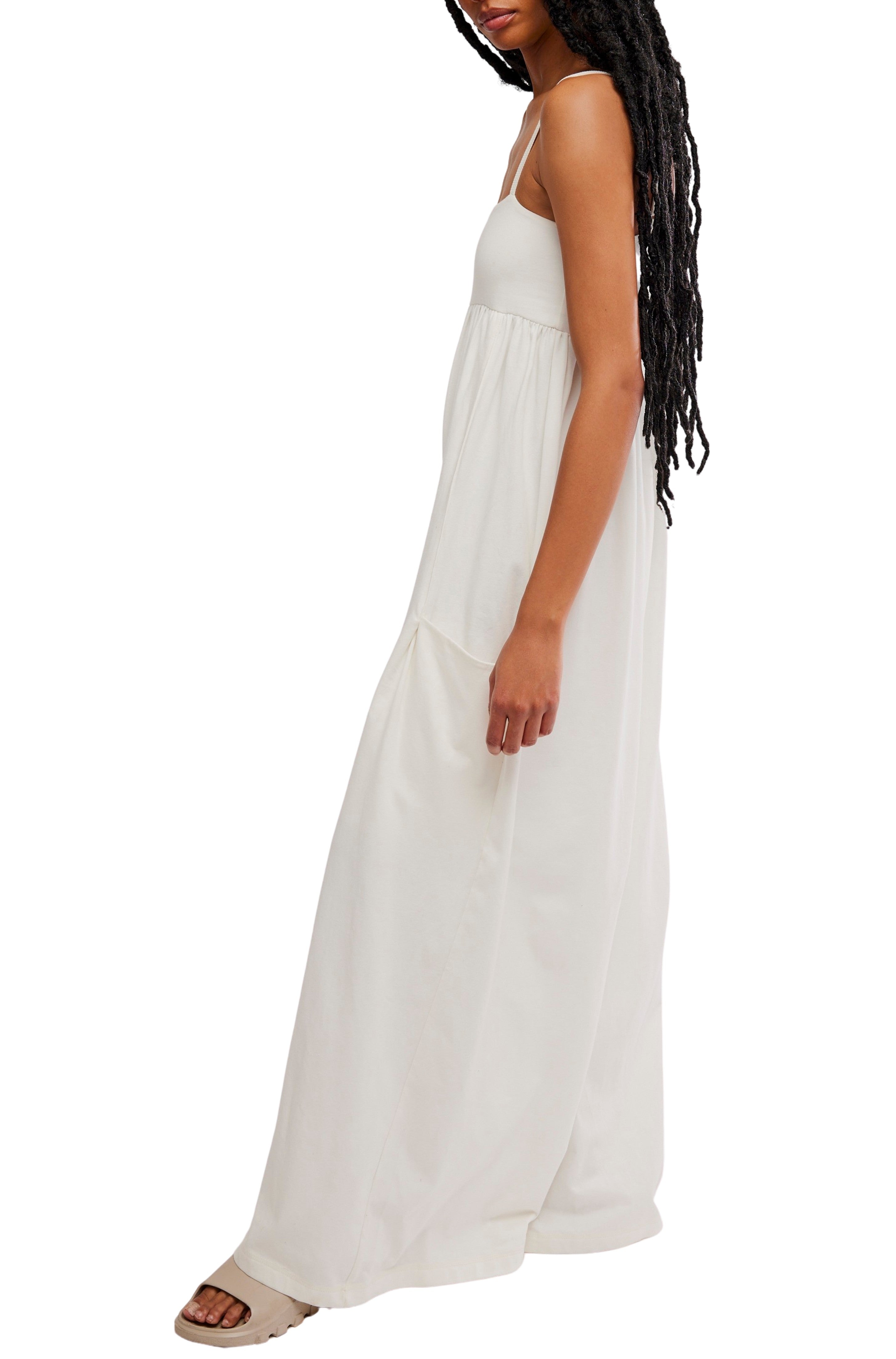 Woman wearing the FP Movement Hot Shot Strappy Jumpsuit, a sleeveless, floor-length white outfit with spaghetti straps and a square neckline, standing sideways against a white background. She has long braided hair and wears sandals.