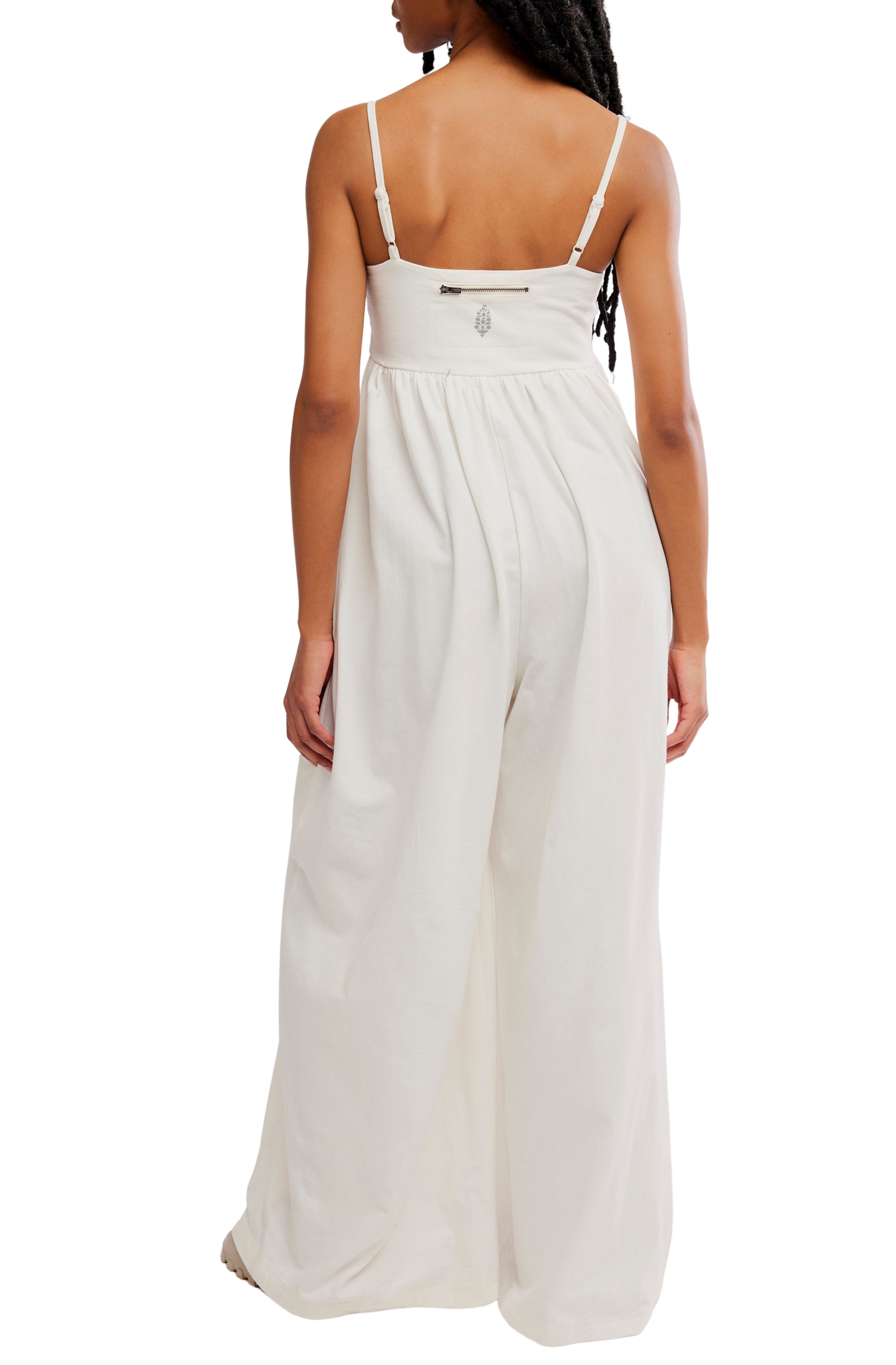 A person with long hair is seen from the back against a white background wearing the FP Movement Hot Shot Strappy Jumpsuit—a sleeveless, wide-leg white style with thin straps and a square neckline.