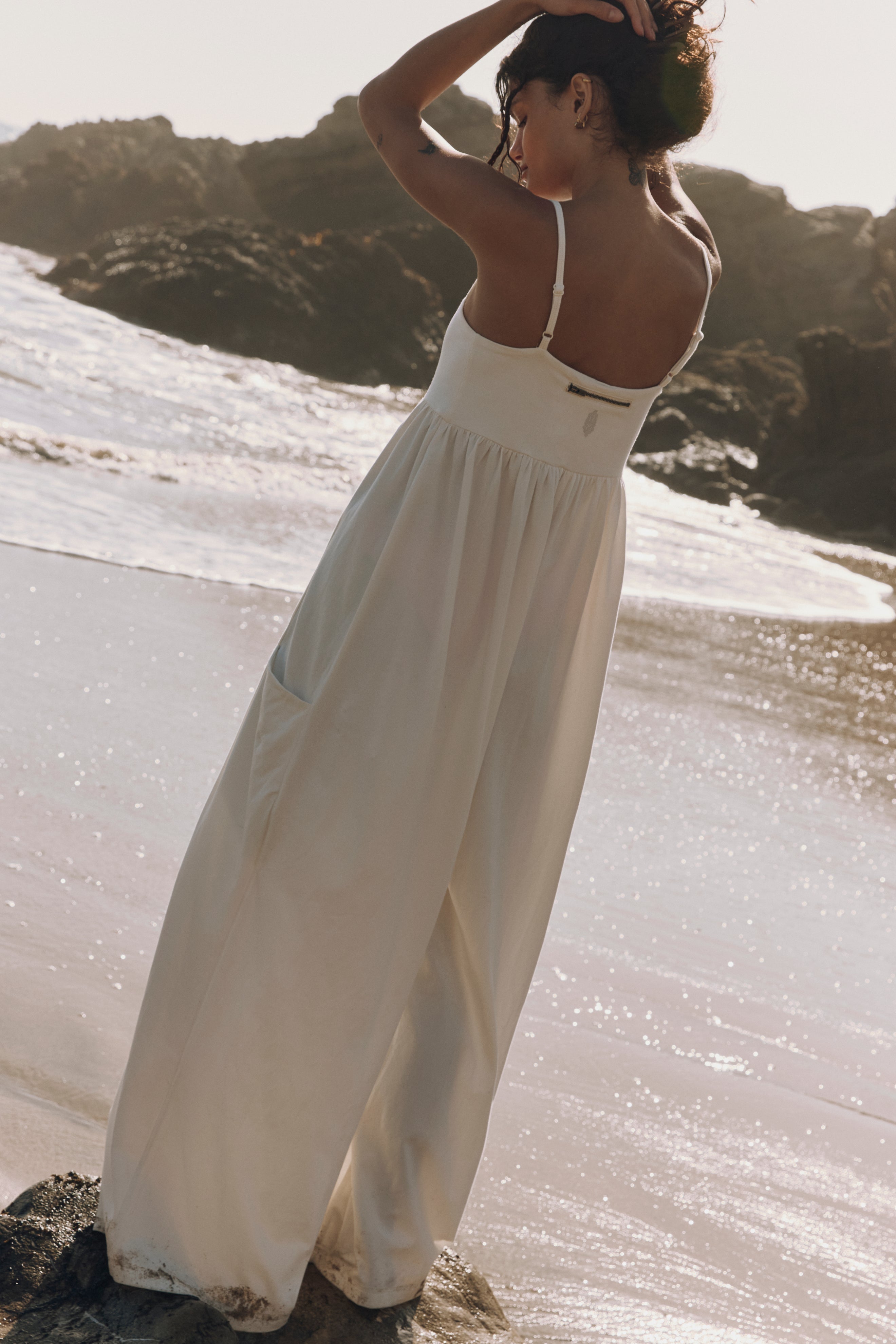 A woman stands barefoot on a rocky beach, facing the ocean in FP Movement's Hot Shot Strappy Jumpsuit, with one hand in her hair as waves and rocks behind her are softly lit by sunlight.