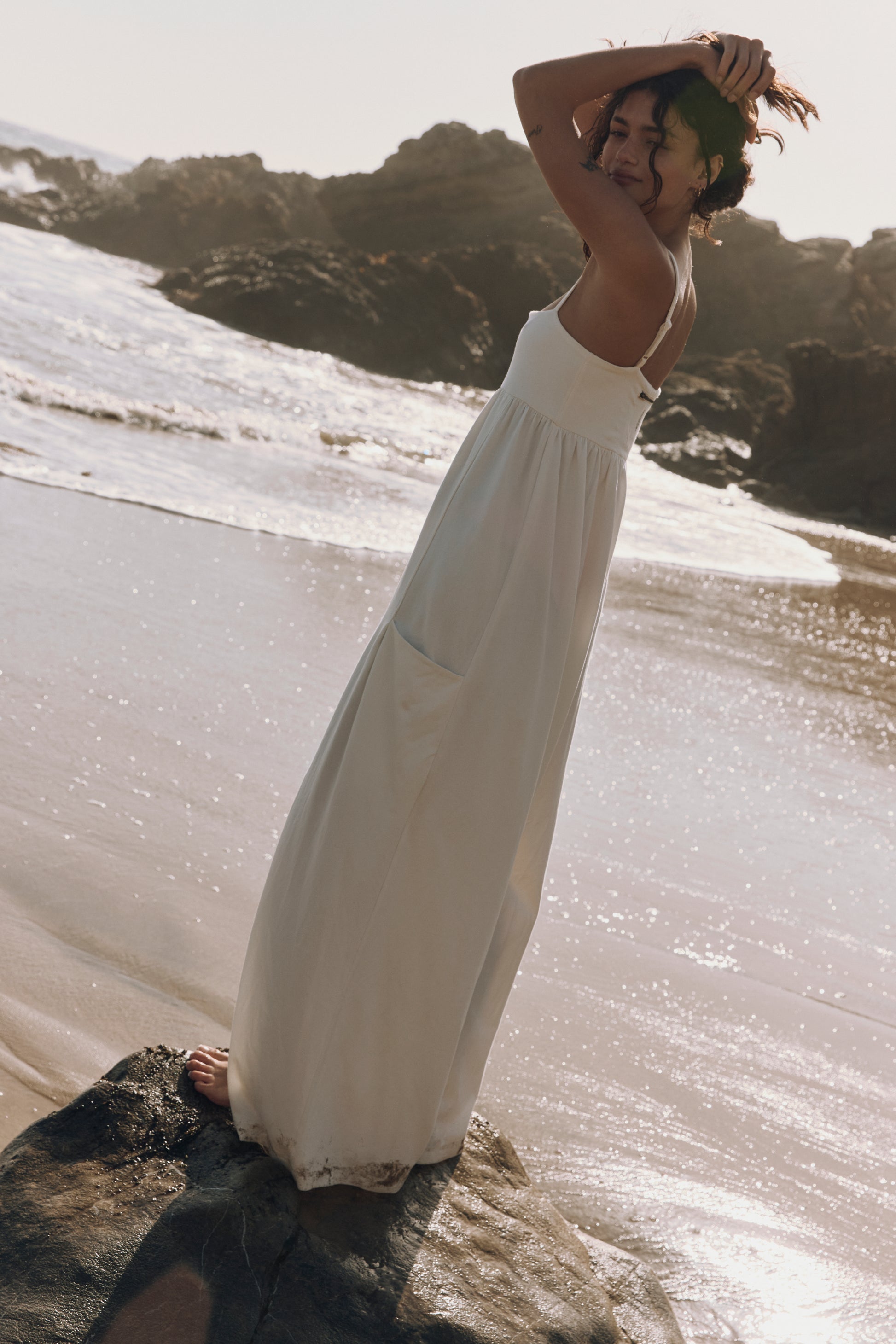 A woman smiles barefoot on a beach rock, wearing the FP Movement Hot Shot Strappy Jumpsuit. She stands with one hand in her hair as sunlight highlights the waves and rocky shoreline behind her.