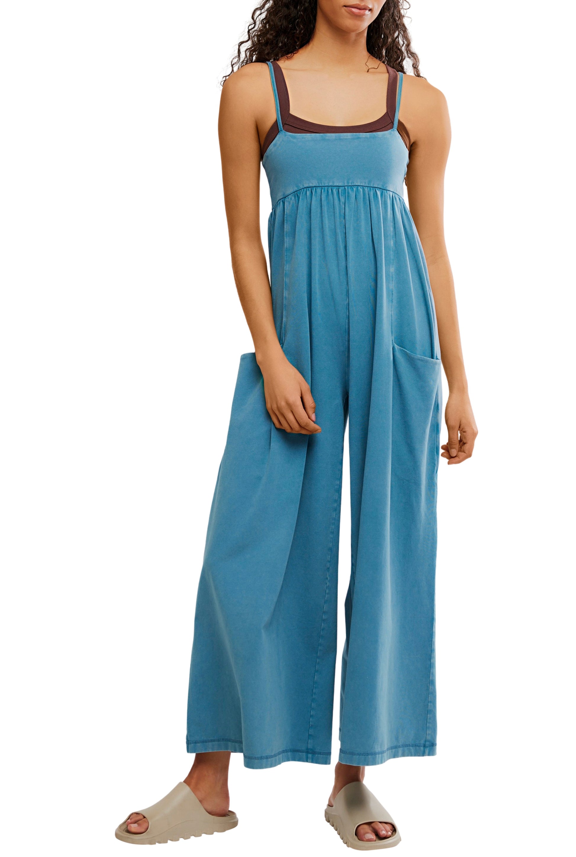Model in the FP Movement Hot Shot Strappy Jumpsuit in blue, featuring large front pockets, styled over a dark brown tank top and light beige slide sandals, standing against a white background.