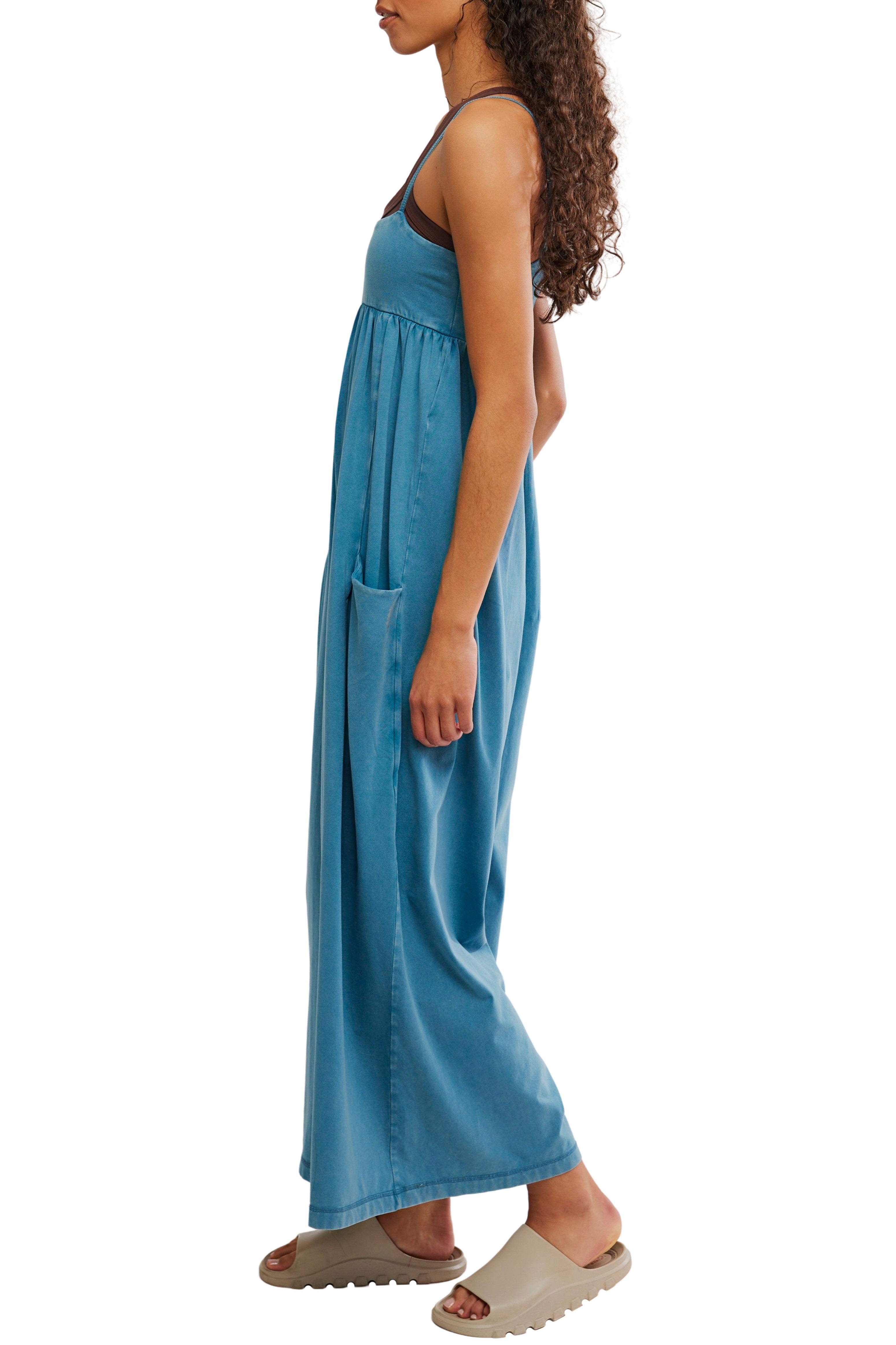 A woman in the FP Movement Hot Shot Strappy Jumpsuit stands in profile, showcasing her curly hair, tan slide sandals, and a casual, relaxed vibe.