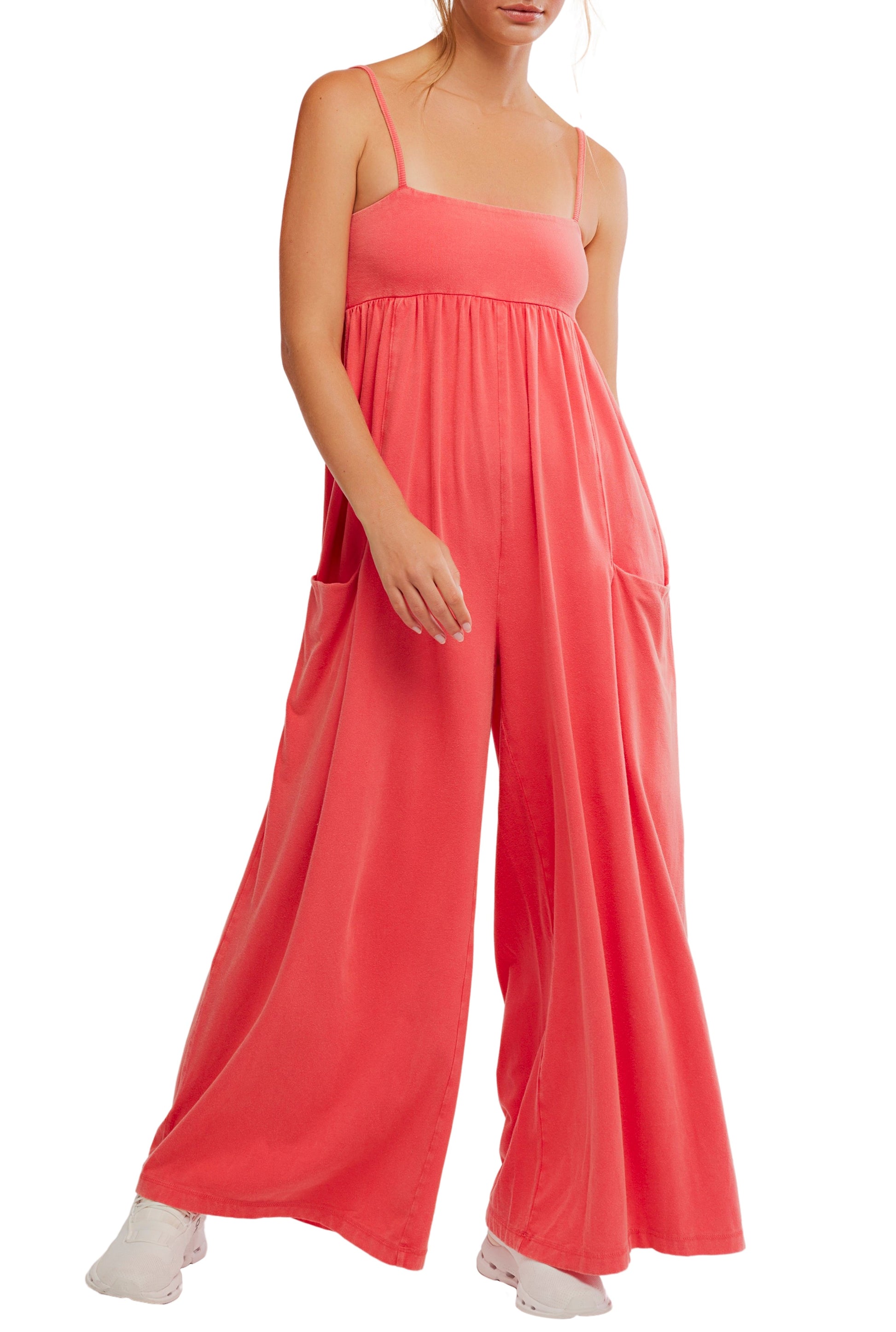 A woman wears the FP Movement Hot Shot Strappy Jumpsuit in coral, featuring a loose fit, spaghetti straps, wide legs, and large pockets. She pairs it with white sneakers and has her hand in one pocket.