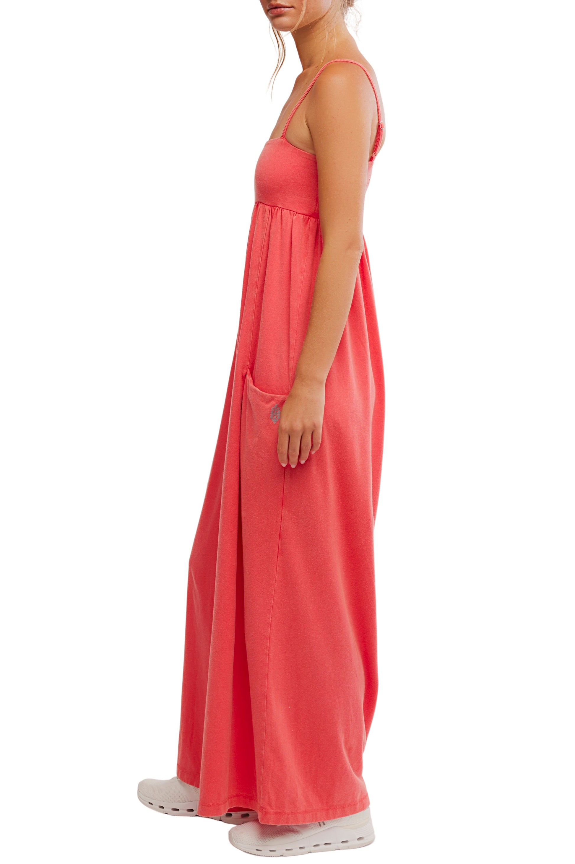 A woman stands in profile wearing the FP Movement Hot Shot Strappy Jumpsuit. Her hair is tied back, and she pairs the jumpsuit with white slip-on shoes. The background is white.