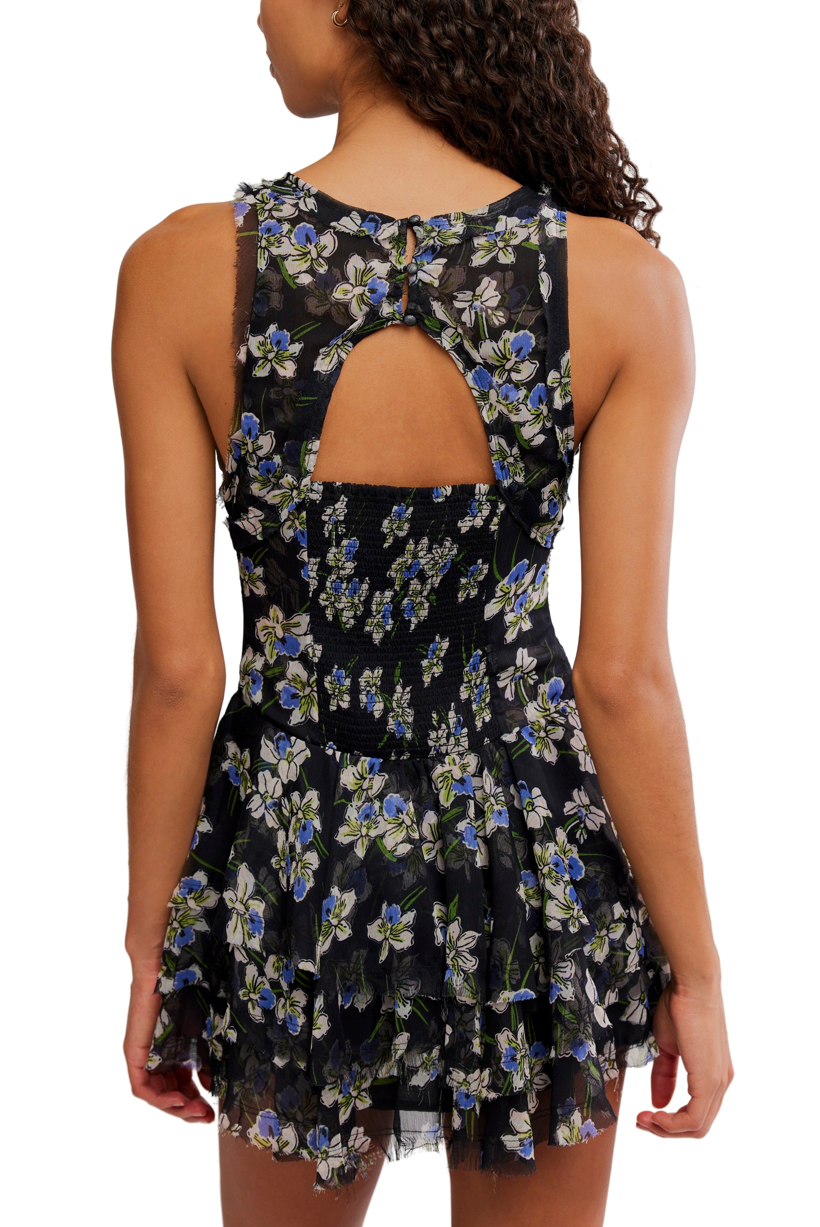 A woman wears the Raven Printed Mini Dress by Free People—a sleeveless black mini with blue and white florals, keyhole cutout, smock back, and a ruffle skirt—standing with her back to the camera.