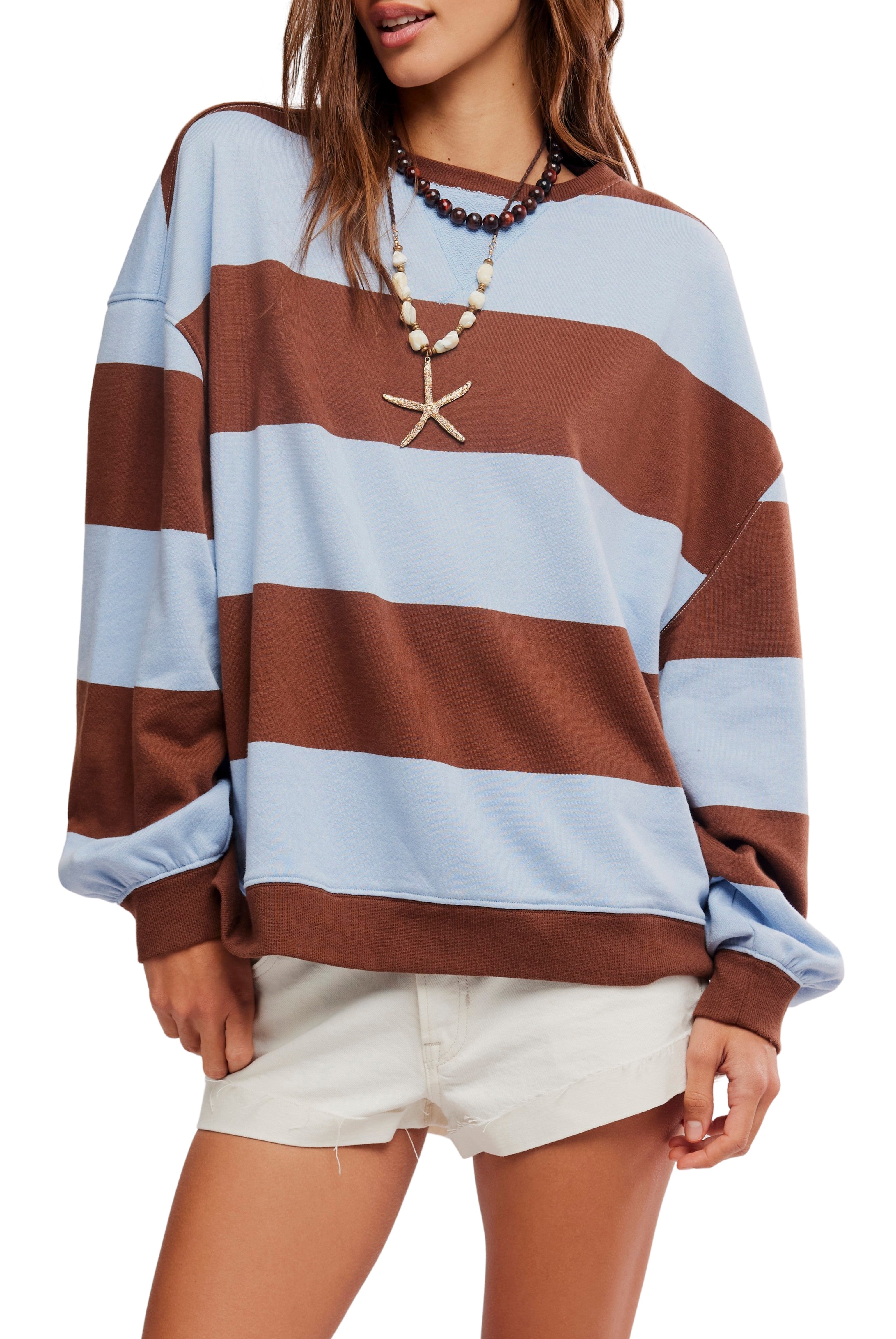 A woman wears Free People’s Classic Stripe Crew Wide sweater with wide brown and light blue stripes, styled with white frayed shorts and shell necklaces for a relaxed, effortless vibe.