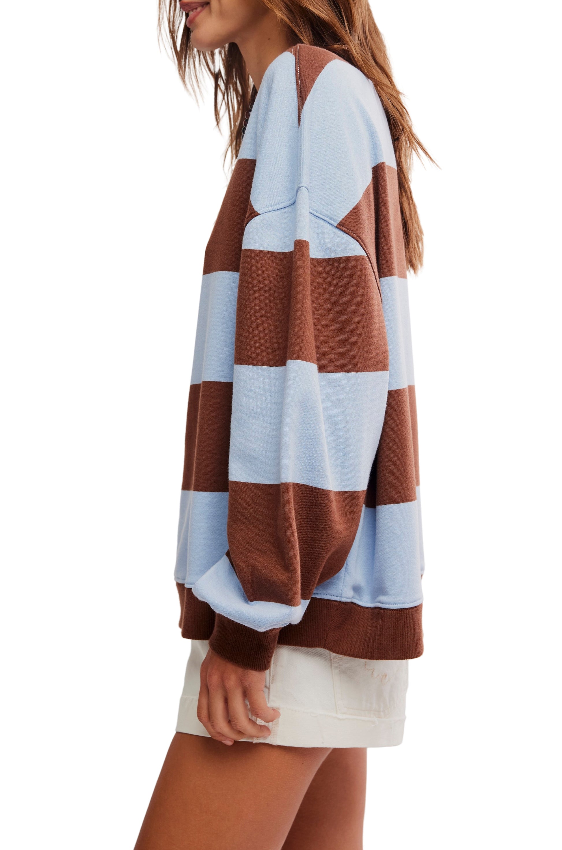 A person wears the Free People Classic Stripe Crew Wide, an oversized light blue and brown striped sweater with a relaxed fit, paired with white shorts. The side-view image is set against a white background.