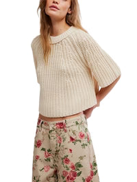 A woman wears the Free People Amy Sweater Tee, a cream short-sleeve knit with ribbed trim, paired with high-waisted beige pants featuring a pink floral print, standing with one arm behind her back.