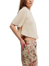 A woman stands in profile wearing the Free People Amy Sweater Tee, a loose cream knit with a crew neckline and ribbed trim, paired with high-waisted beige pants featuring a pink floral print. Her right hand rests in her pocket, face partly out of frame.