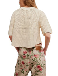 A person shown from the back wears the Free People Amy Sweater Tee in cream, a chunky knit with ribbed trim, paired with high-waisted beige pants featuring pink and green floral patterns.